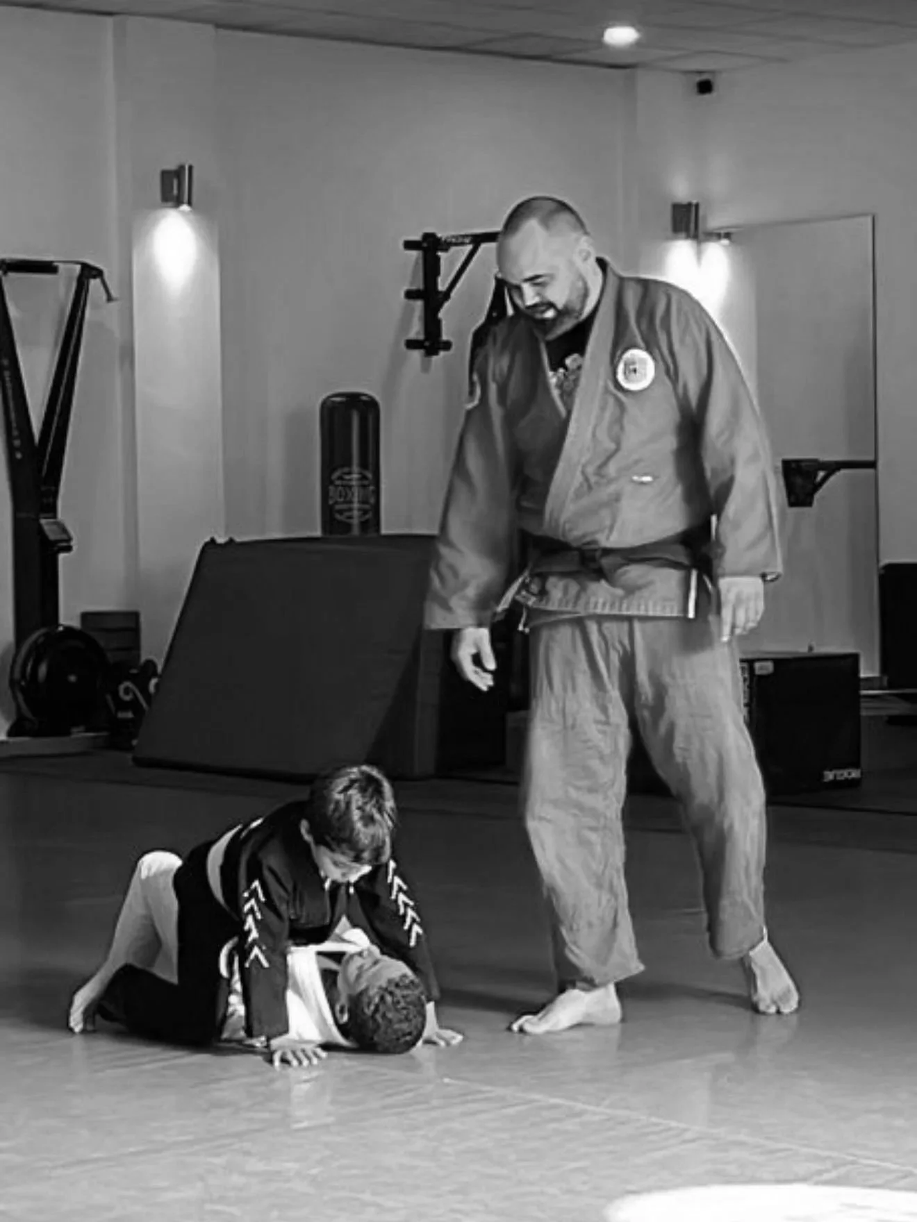 A martial arts instructor in a gi demonstrating a technique to two young students on a training mat indoors at Guild BJJ academy in Wandsworth, London