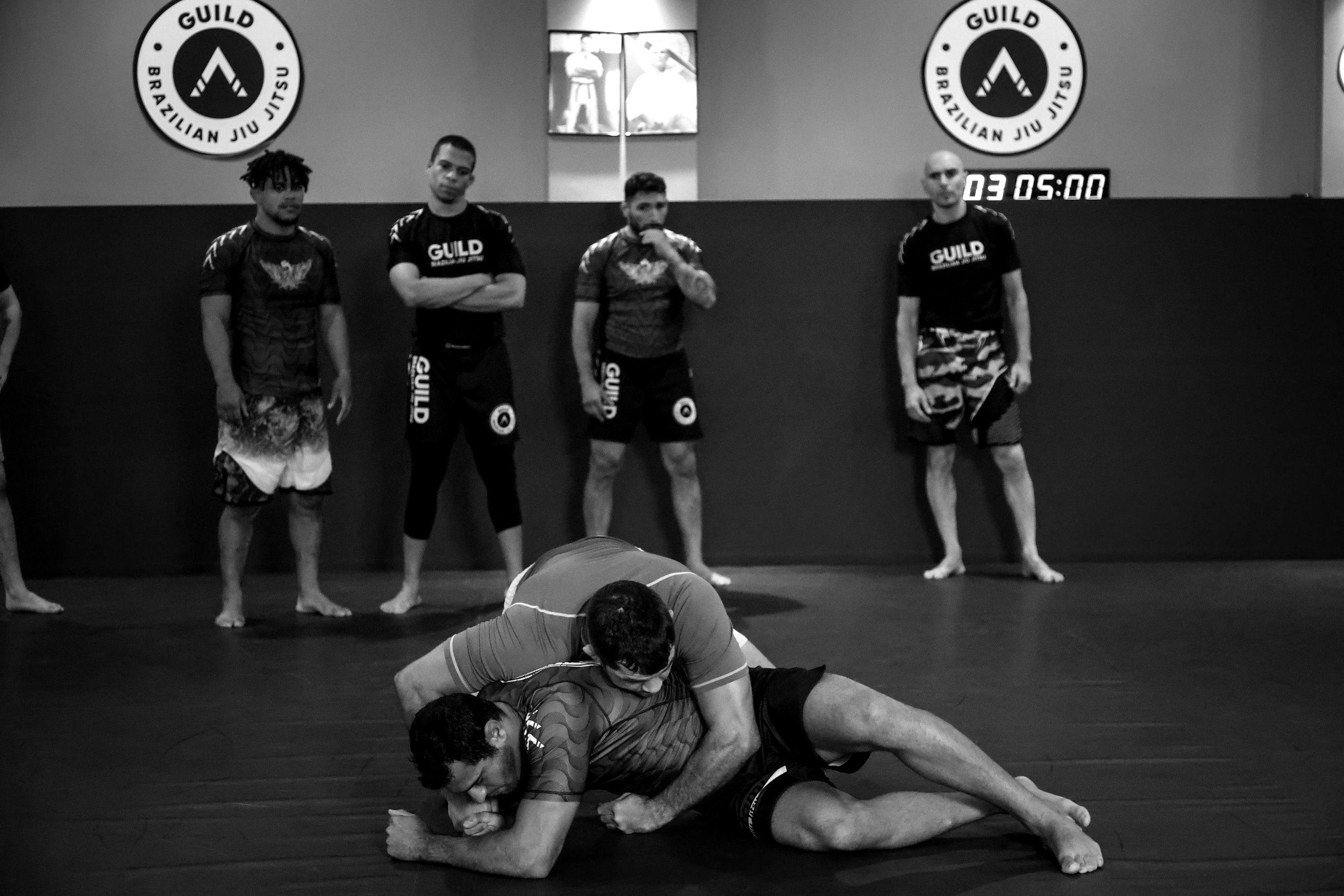 A group of people practicing Brazilian jiu-jitsu in a gym, with four individuals standing against the wall and two wrestling on the mat in the foreground.