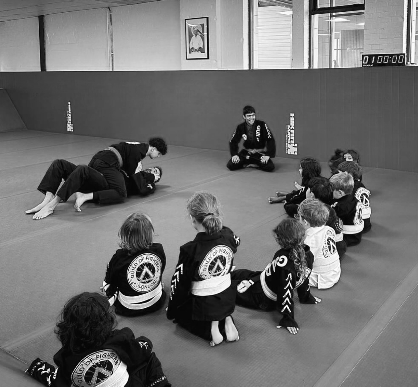 Children dressed in Brazilian Jiu-Jitsu uniforms sitting on a mat in a martial arts dojo, listening to an instructor demonstrating a technique while another instructor performs a move with a student.