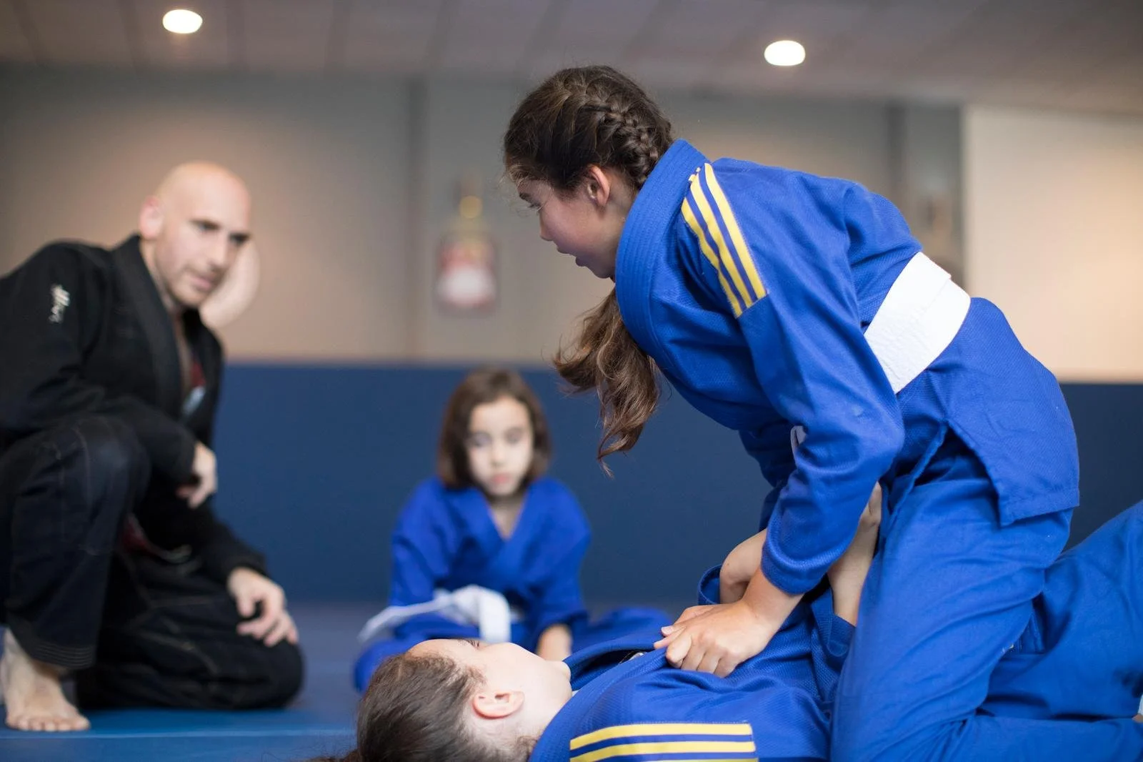 Two young girls in martial arts uniforms practicing Brazilian Jiu-Jitsu kids classes on mats, with an instructor and another girl in black martial arts uniform observing and coaching.