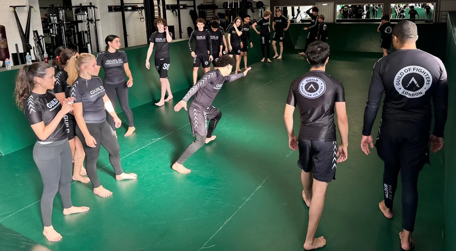 Group of people practicing Brazilian Jiu-Jitsu in a gym. They are wearing matching black uniforms with the words 'GUILD' on them, inside a room with padded green floors and mirrors in Wandsworth, London.