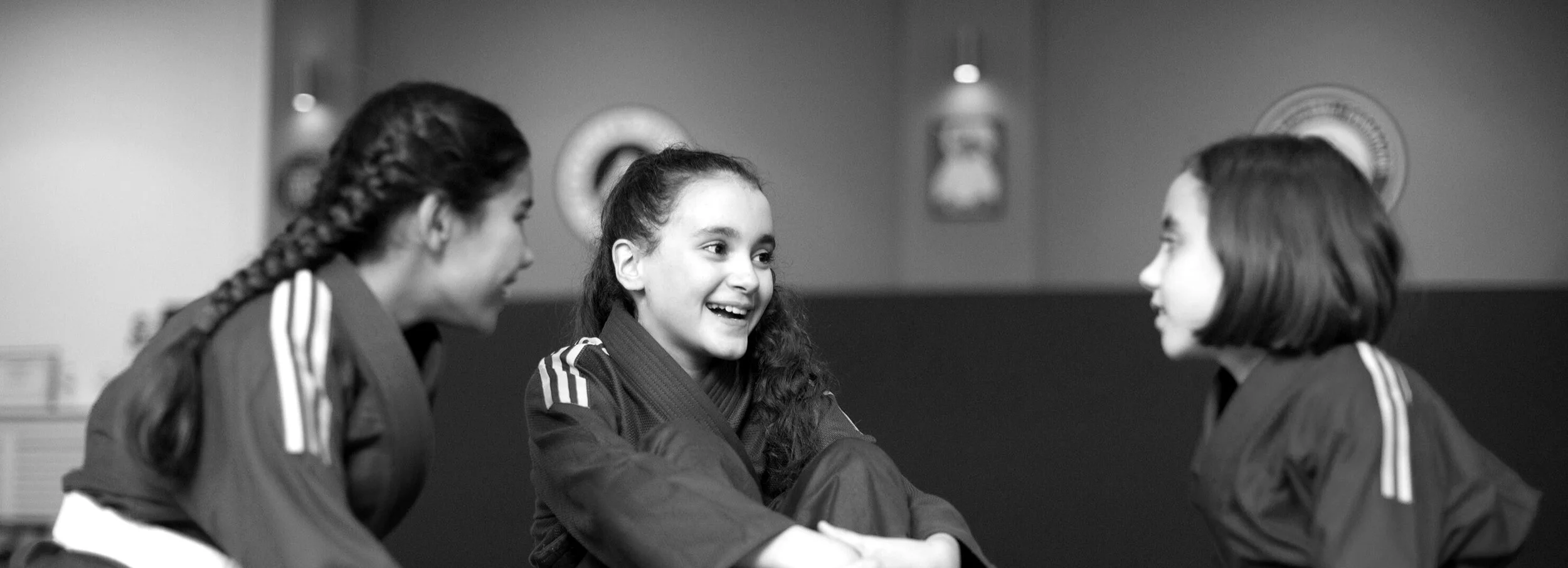 Three young women in martial arts uniforms engaging in a friendly conversation or handshake, smiling, in a training facility that offers womens only BJJ classes