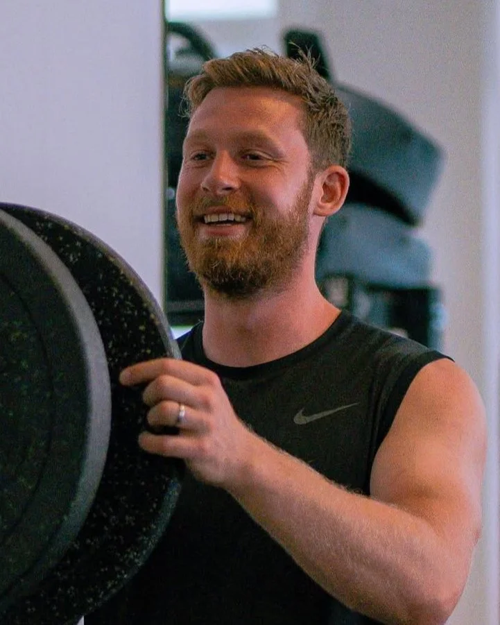A man with a beard and short hair smiling while lifting a weight in a gym in SW18