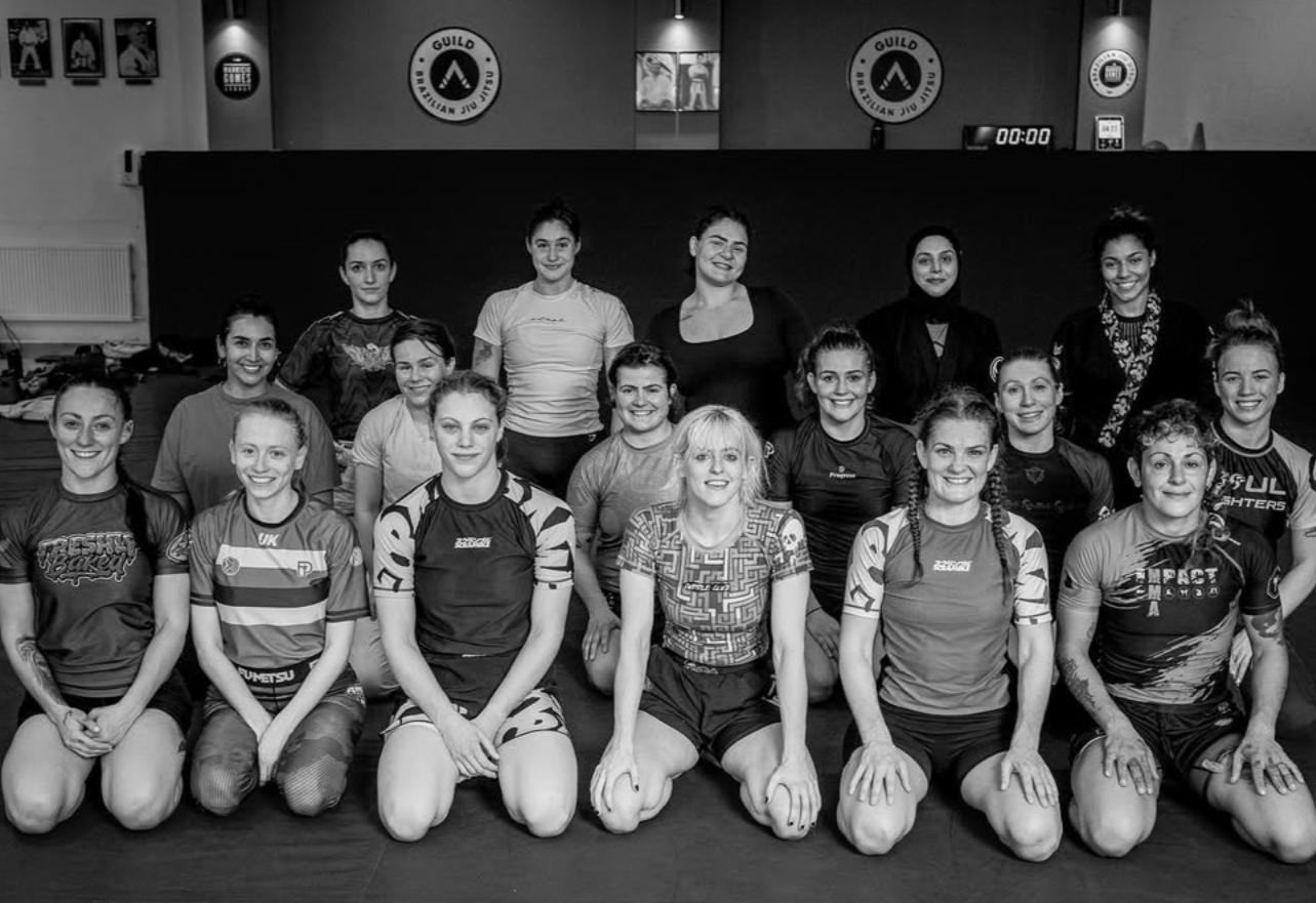 Group photo of female athletes in a mixed martial arts gym, sitting and standing on a mat, with training logos and posters on the wall behind them.