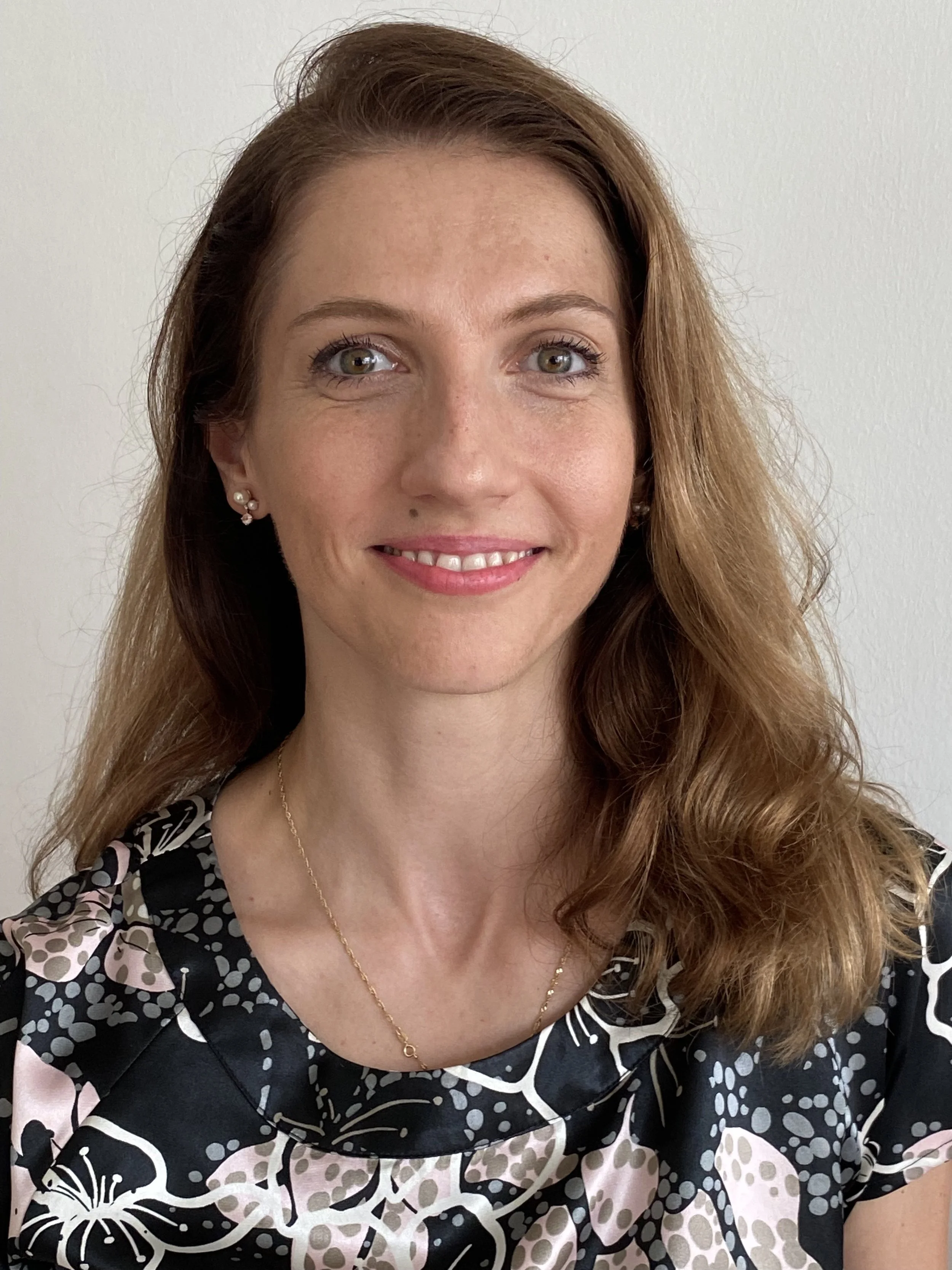 A woman with long, wavy, auburn hair smiling at the camera, wearing pearl earrings, a gold necklace, and a patterned blouse with black, pink, and white colors, standing against a plain white wall.