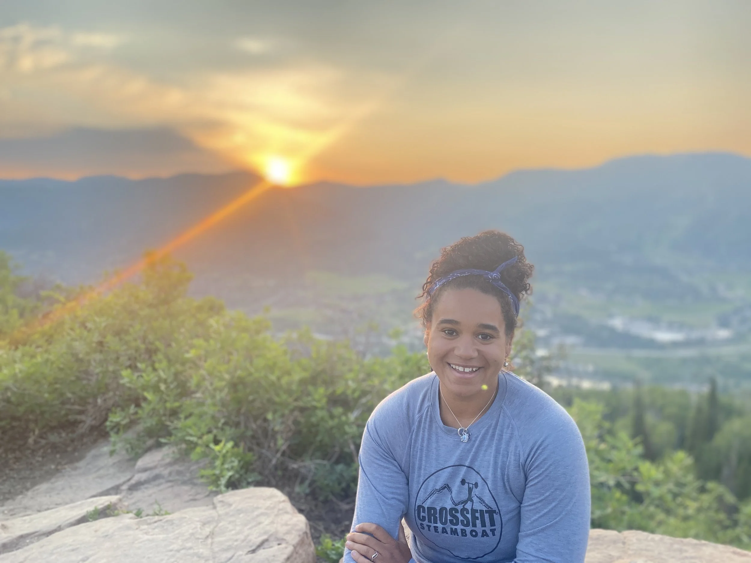 A smiling woman with curly hair tied back in a headband, wearing a blue t-shirt with 'CROSSFIT STEAMBOAT' logo, sitting outdoors at sunset with mountains and greenery in the background.