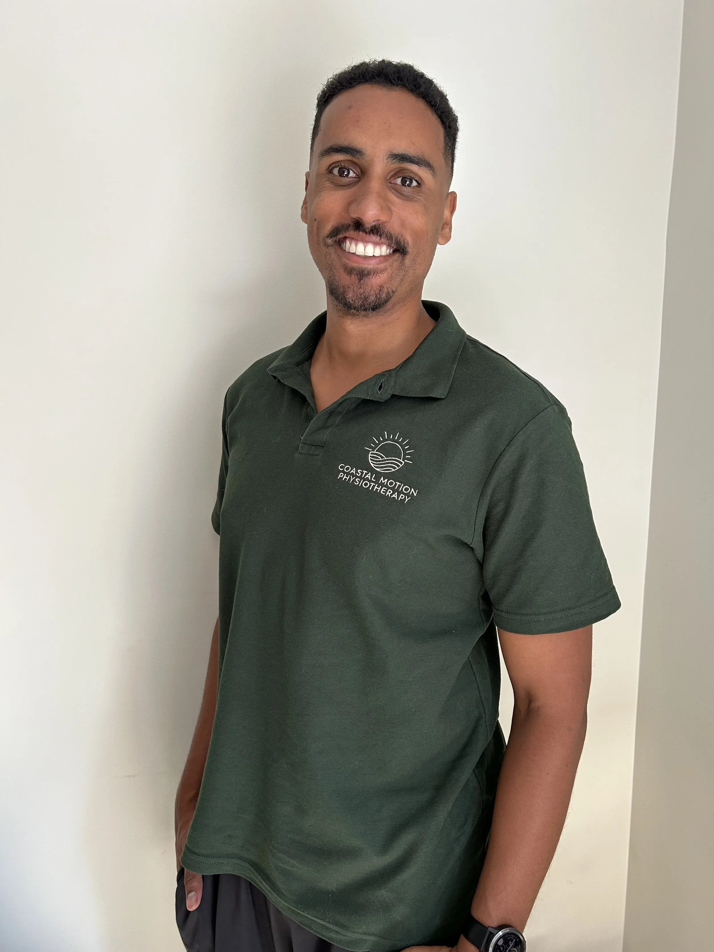 A smiling man with short, curly black hair and a goatee, wearing a dark green polo shirt with a "Coastal Motion Physiotherapy" logo, standing against a white wall.