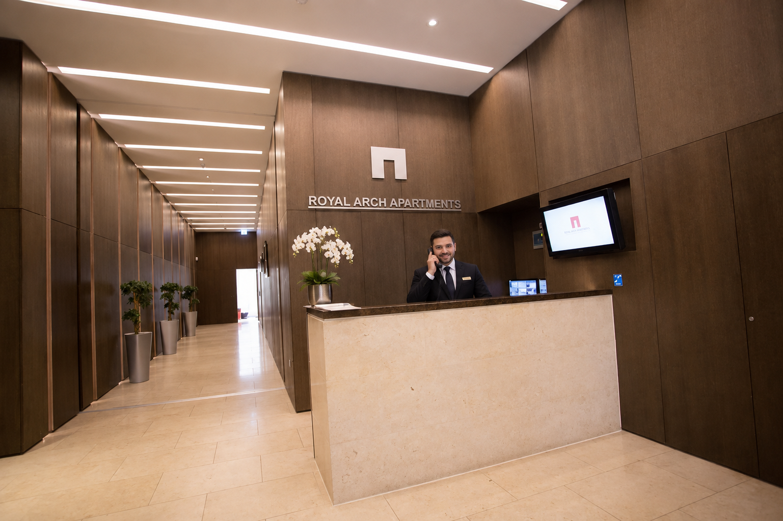Reception area of Royal Arch Apartments with a smiling man on the phone behind the counter, a potted orchid on the desk, wooden wall panels, a wall-mounted screen, and potted plants in silver containers along the hallway.