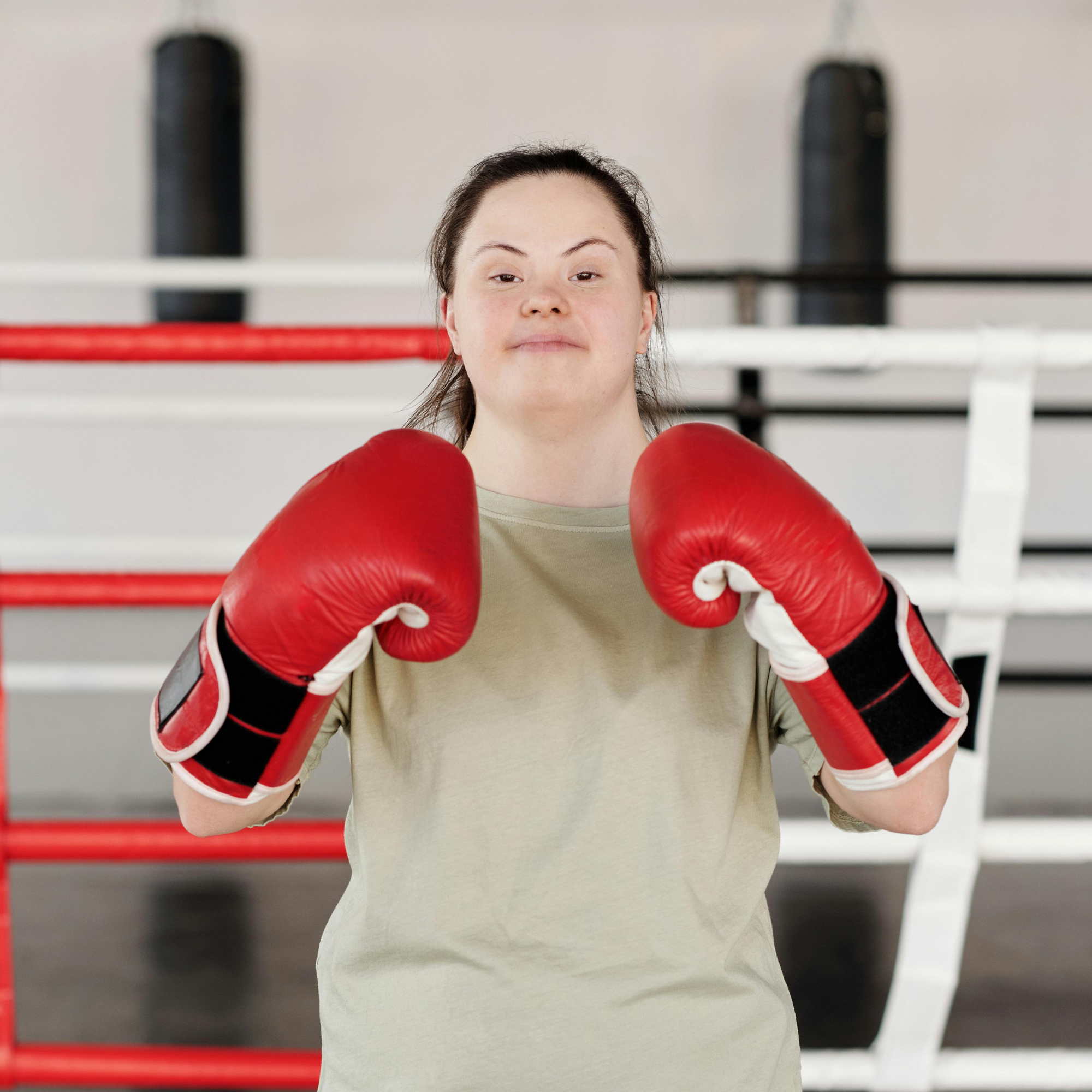 A woman with dark hair in a ponytail wearing red boxing gloves and a beige shirt, standing inside a boxing ring with white ropes and black padding.