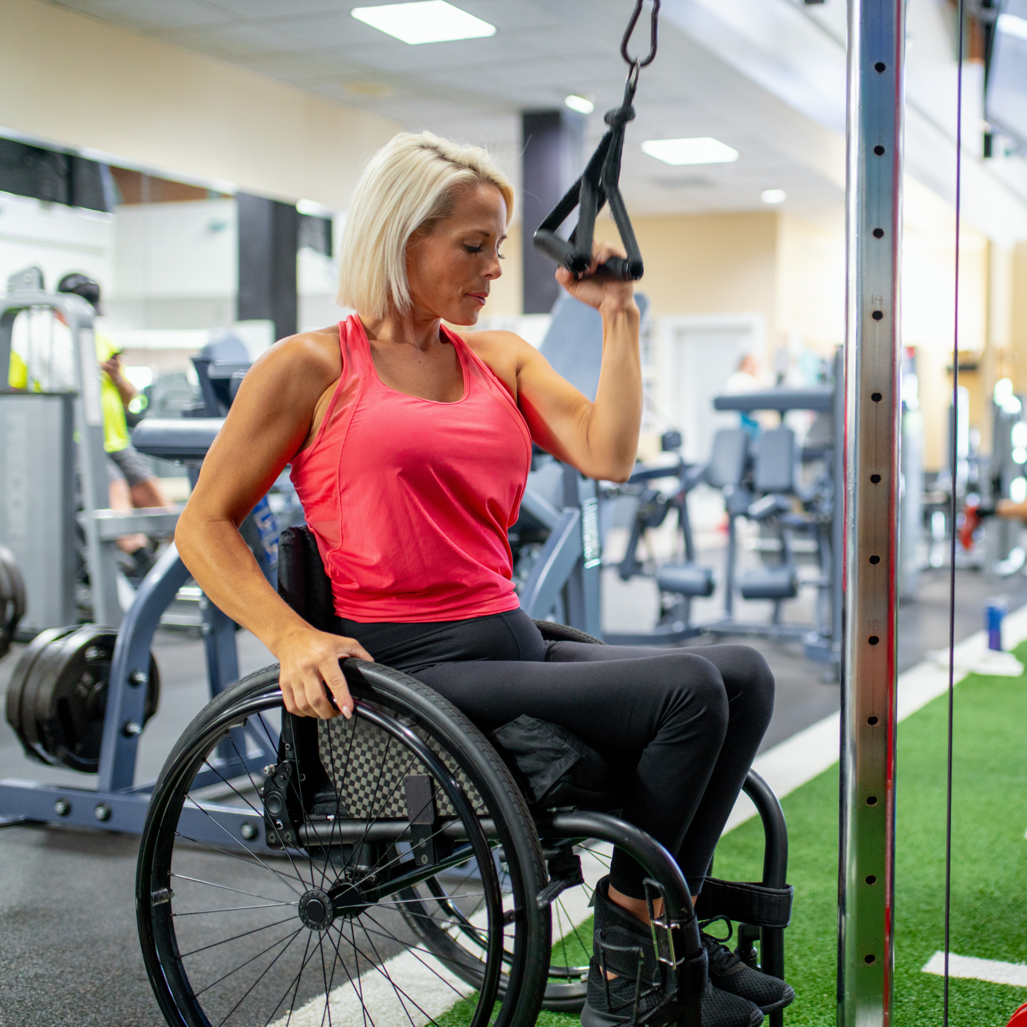 A woman in a pink sports top and black pants, sitting in a wheelchair at the gym, holding her wheelchair armrest with her right hand, and adjusting a pulley in exercise equipment.