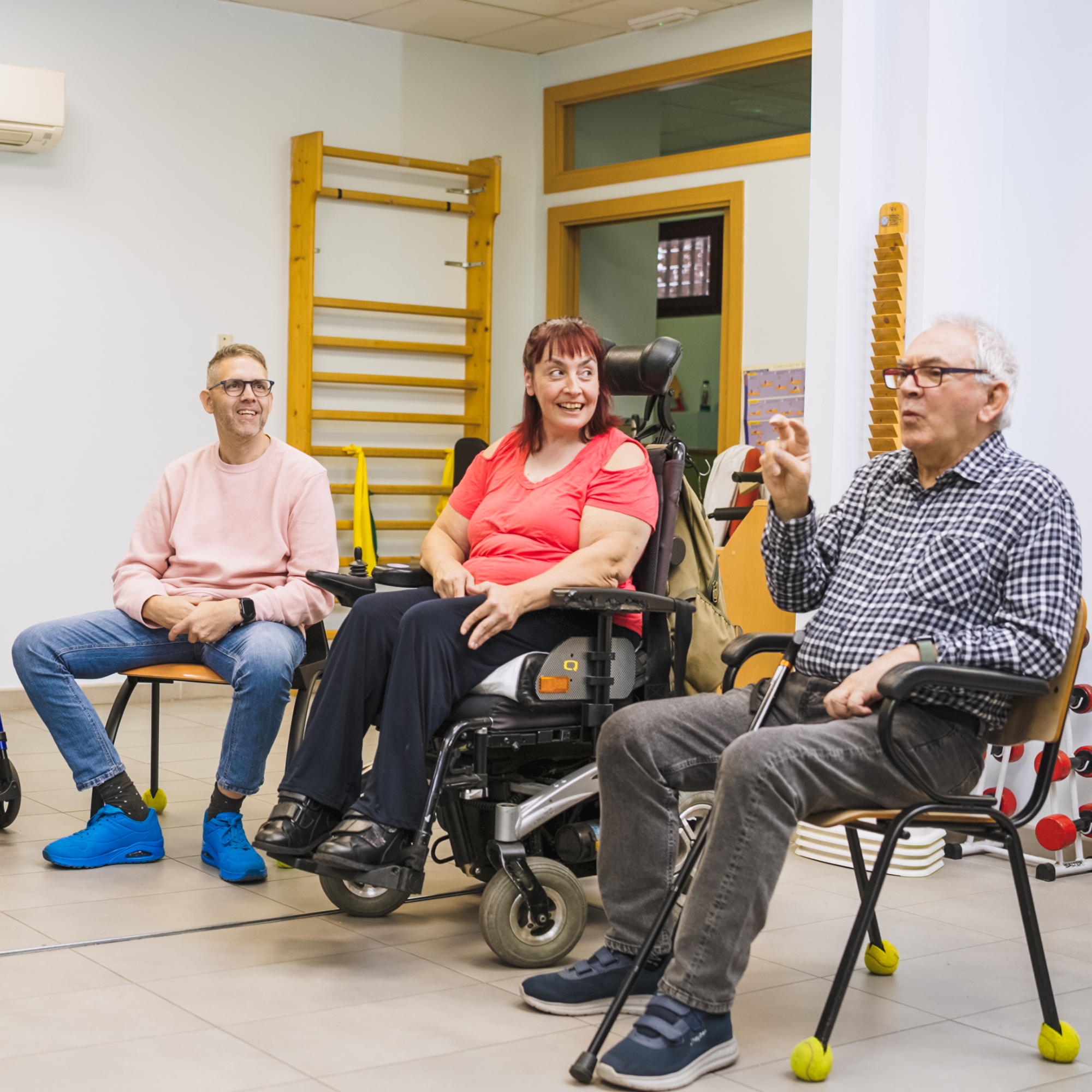 Group of people, some in wheelchairs, in a room with white walls, wooden gym equipment, and a window.