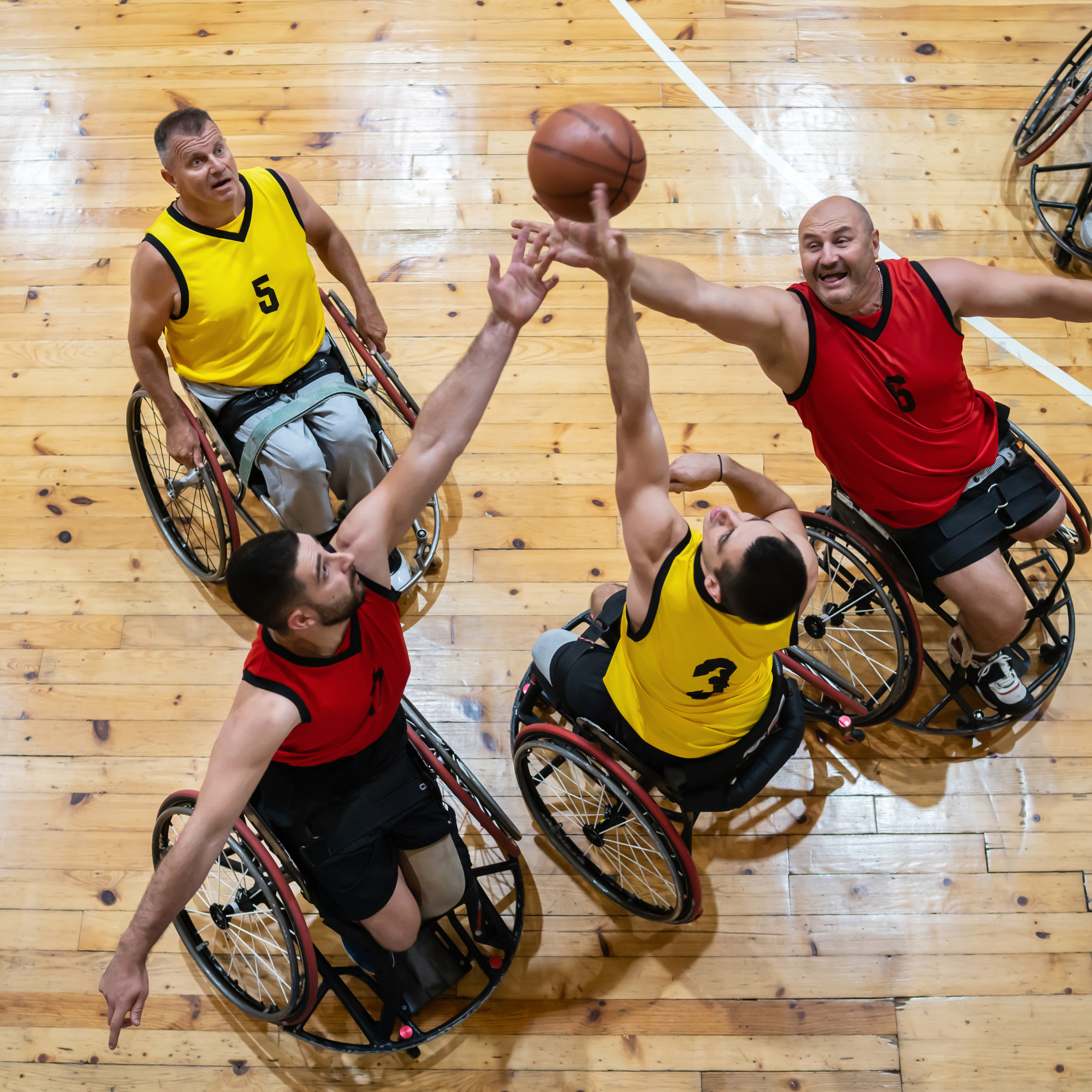 Wheelchair basketball players in action, reaching for the basketball during a game on a polished wooden court.