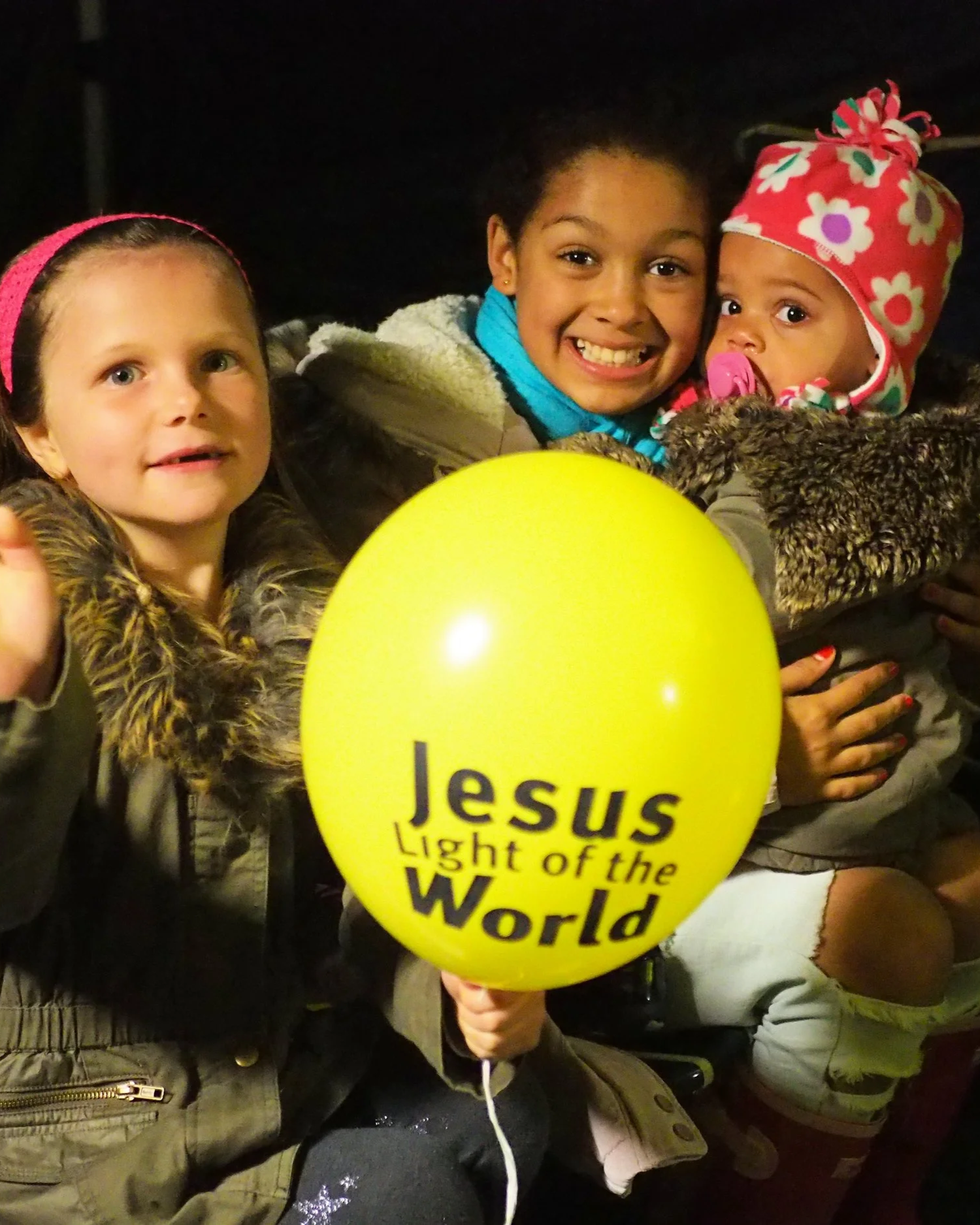 Group of children smiling at nighttime, one holding a yellow balloon with the words 'Jesus Light of the World.'
