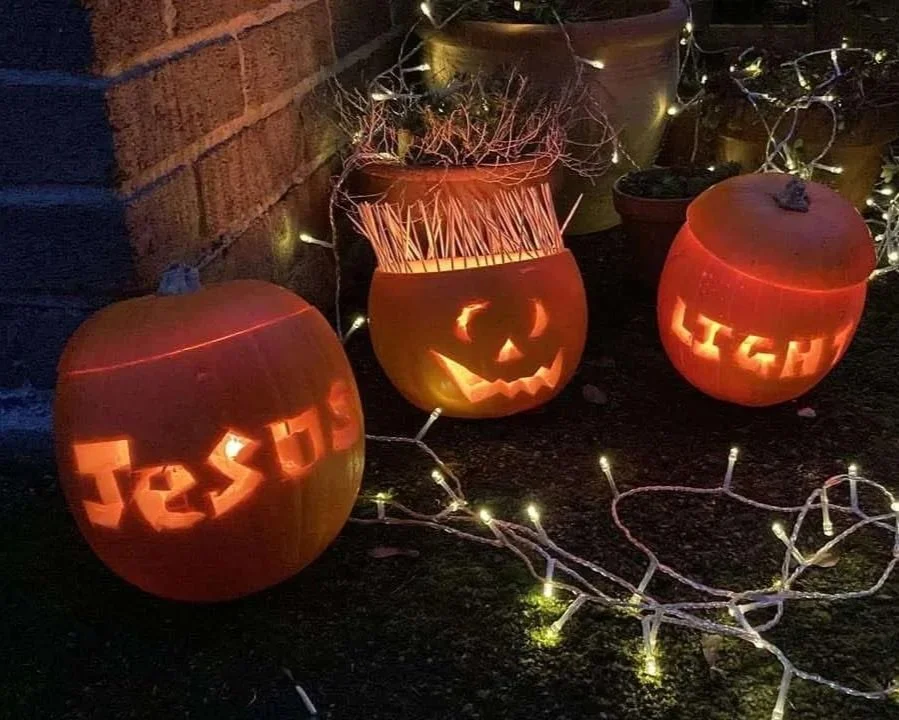 Three carved pumpkins with Halloween-themed designs, surrounded by string lights, on a dark outdoor surface near a brick wall and potted plants.