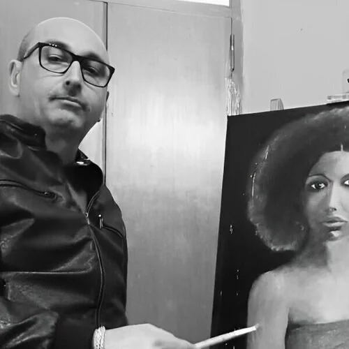 Un uomo con occhiali e giacca di pelle nero che tiene un pennello, accanto a un quadro di una donna con capelli afro e abito nero.
