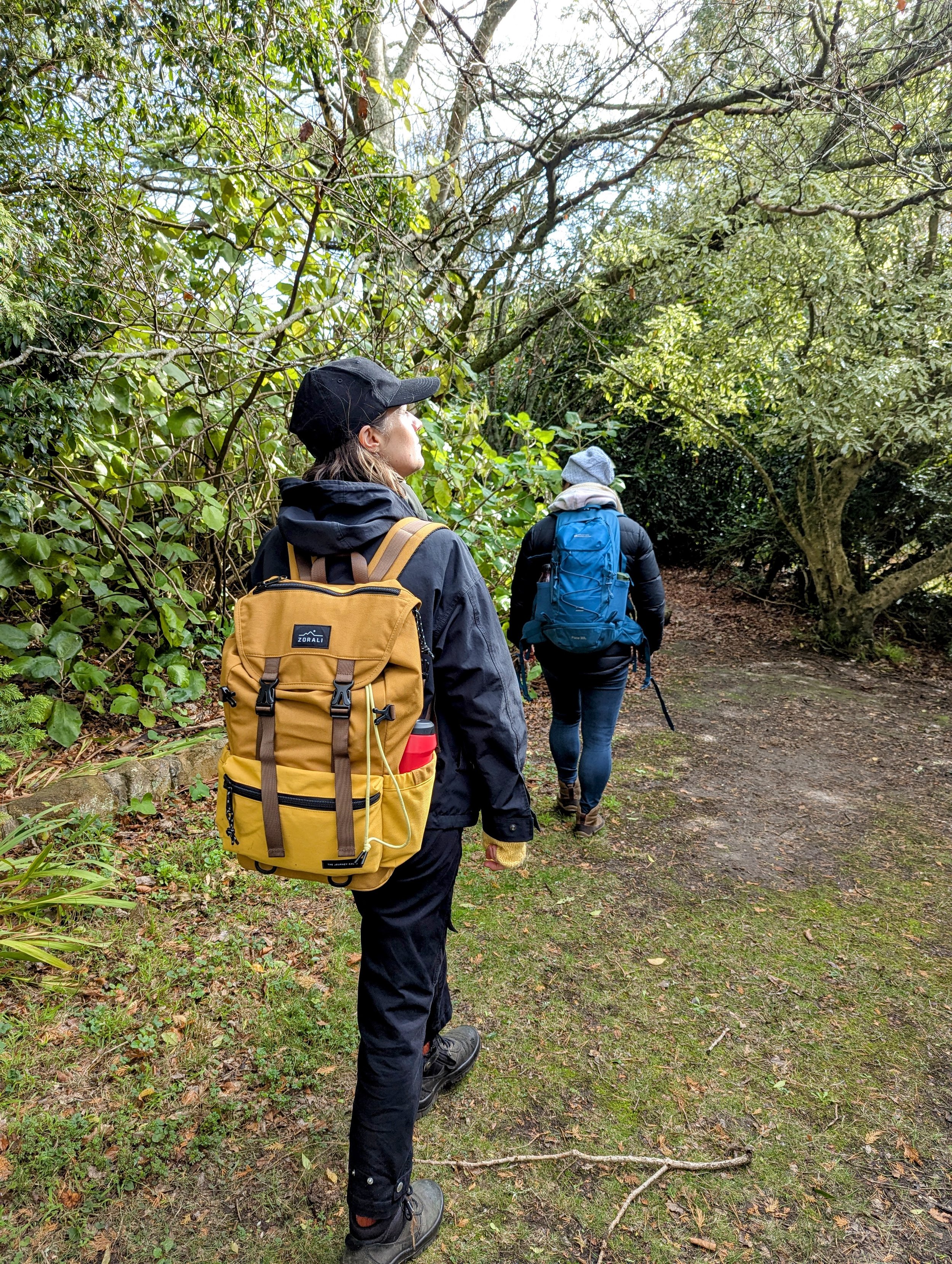 Two people walking on a wooded trail, surrounded by green trees and foliage, carrying backpacks and dressed in outdoor clothing suitable for hiking.