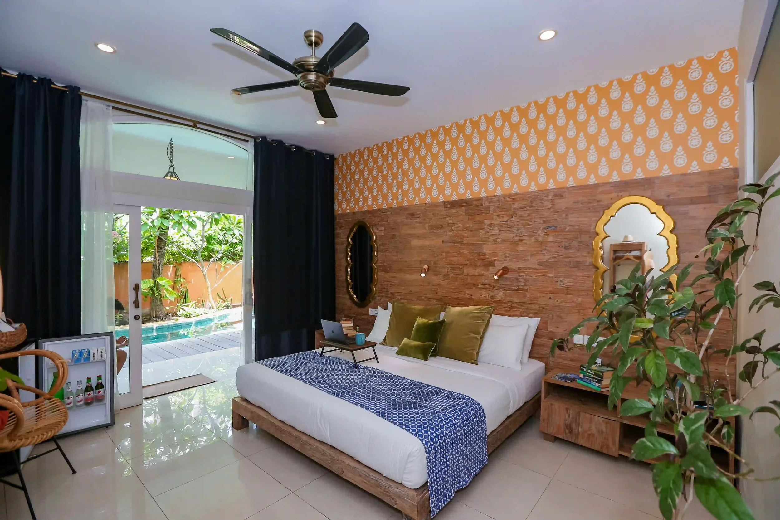A bedroom with a wooden bed frame and a white mattress, decorated with green and white pillows, and a blue patterned blanket. The bedroom has a wooden side table with books and a lamp, a decorative mirror, and a large plant. It features a brick accent wall behind the bed, orange wallpaper with a white pattern on the upper part of the wall, and a window with dark curtains revealing a view of a patio and garden area with trees and a pool outside.