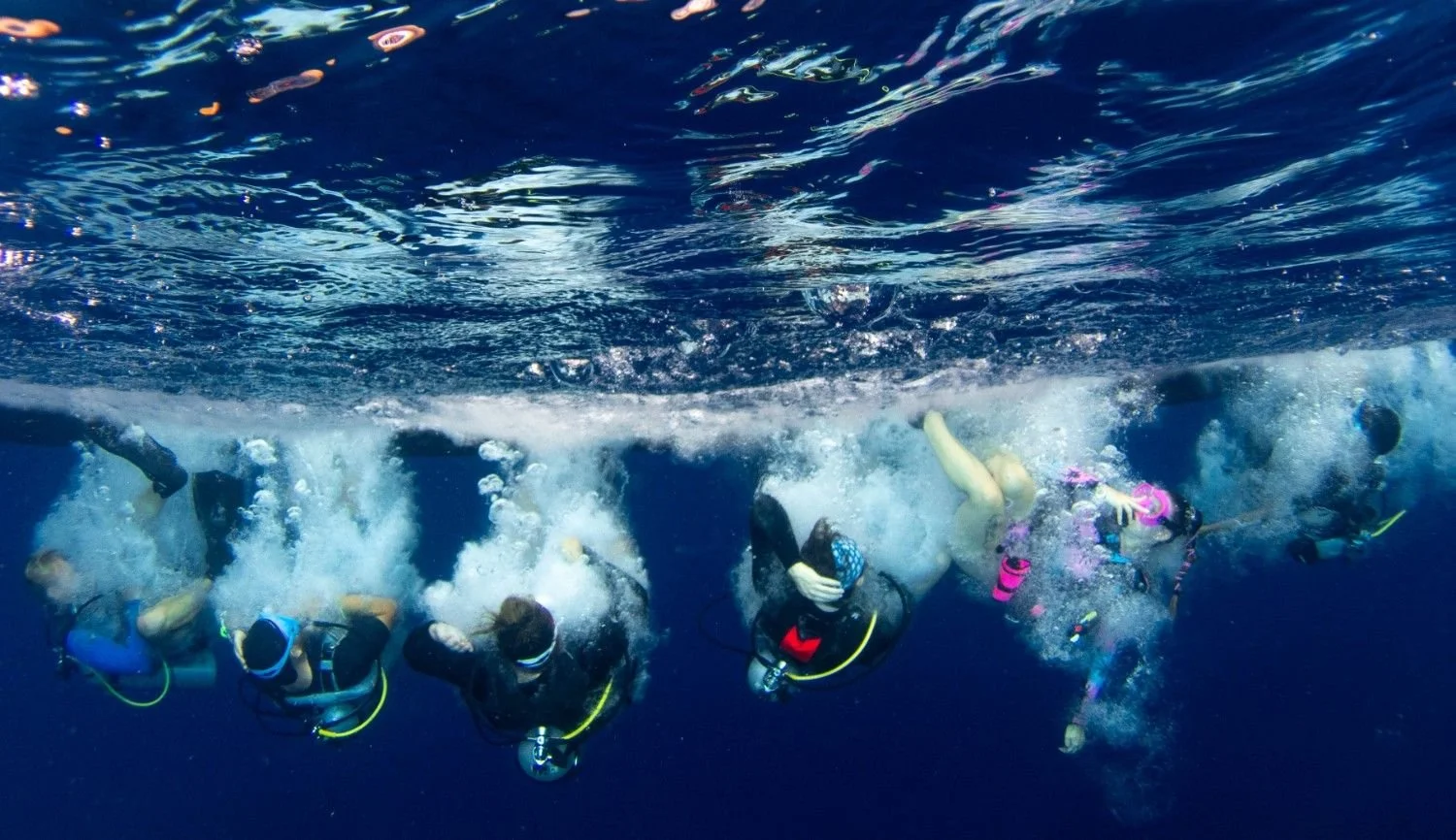 A group of scuba divers entering the water together, with bubbles rising around them beneath the surface.