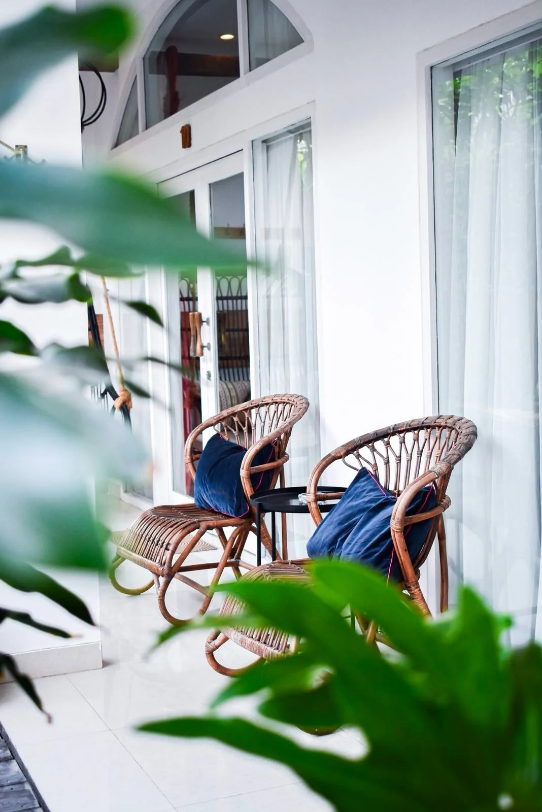 Two wicker chairs with blue cushions on a balcony next to sliding glass doors, framed by green leaves in the foreground.