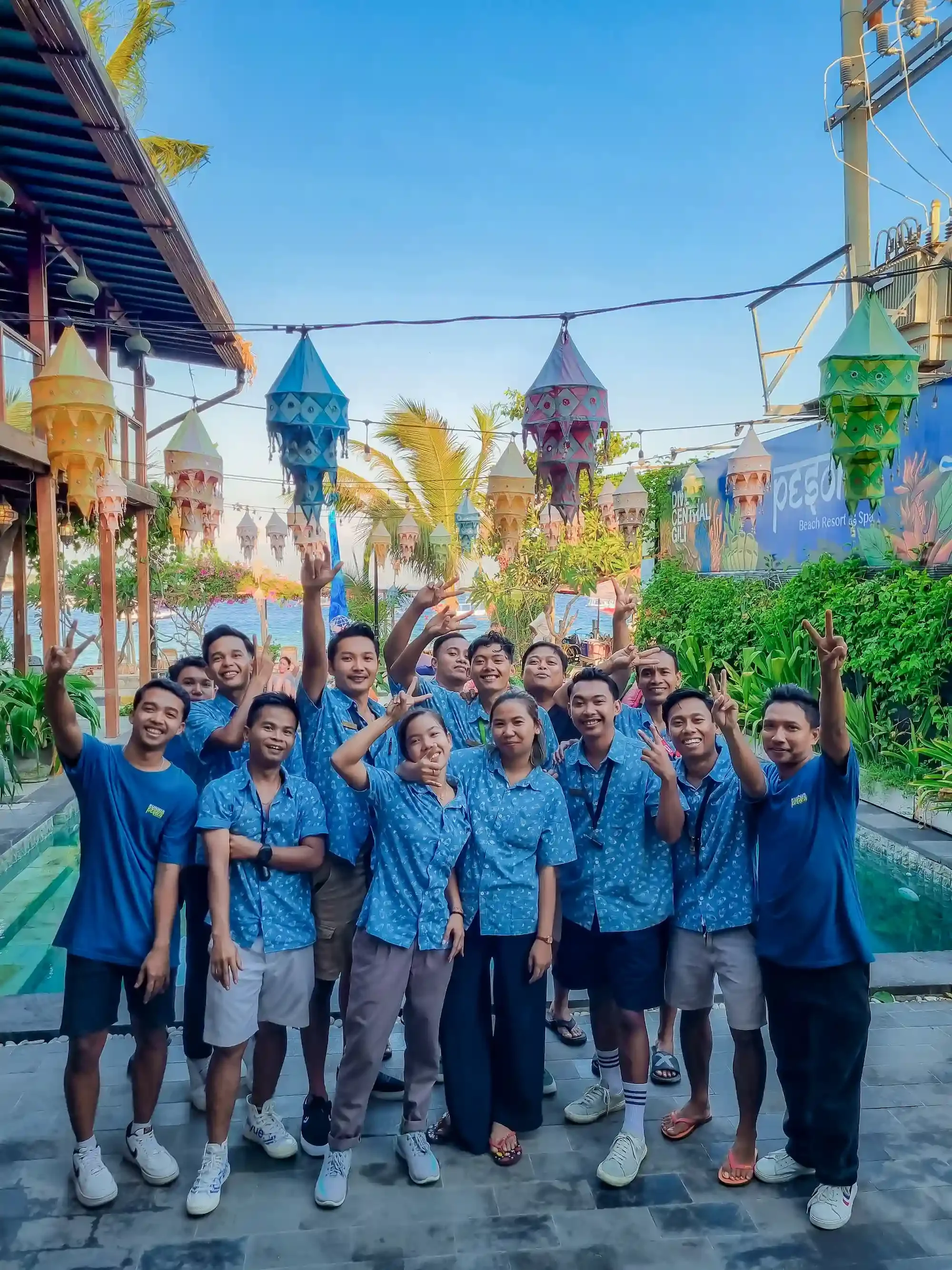 A group of people wearing matching blue shirts with white shorts or pants, posing for a photo outdoors under colorful lanterns on a sunny day. They are smiling and making peace signs, surrounded by lush green plants, and there is the ocean and clear blue sky in the background.