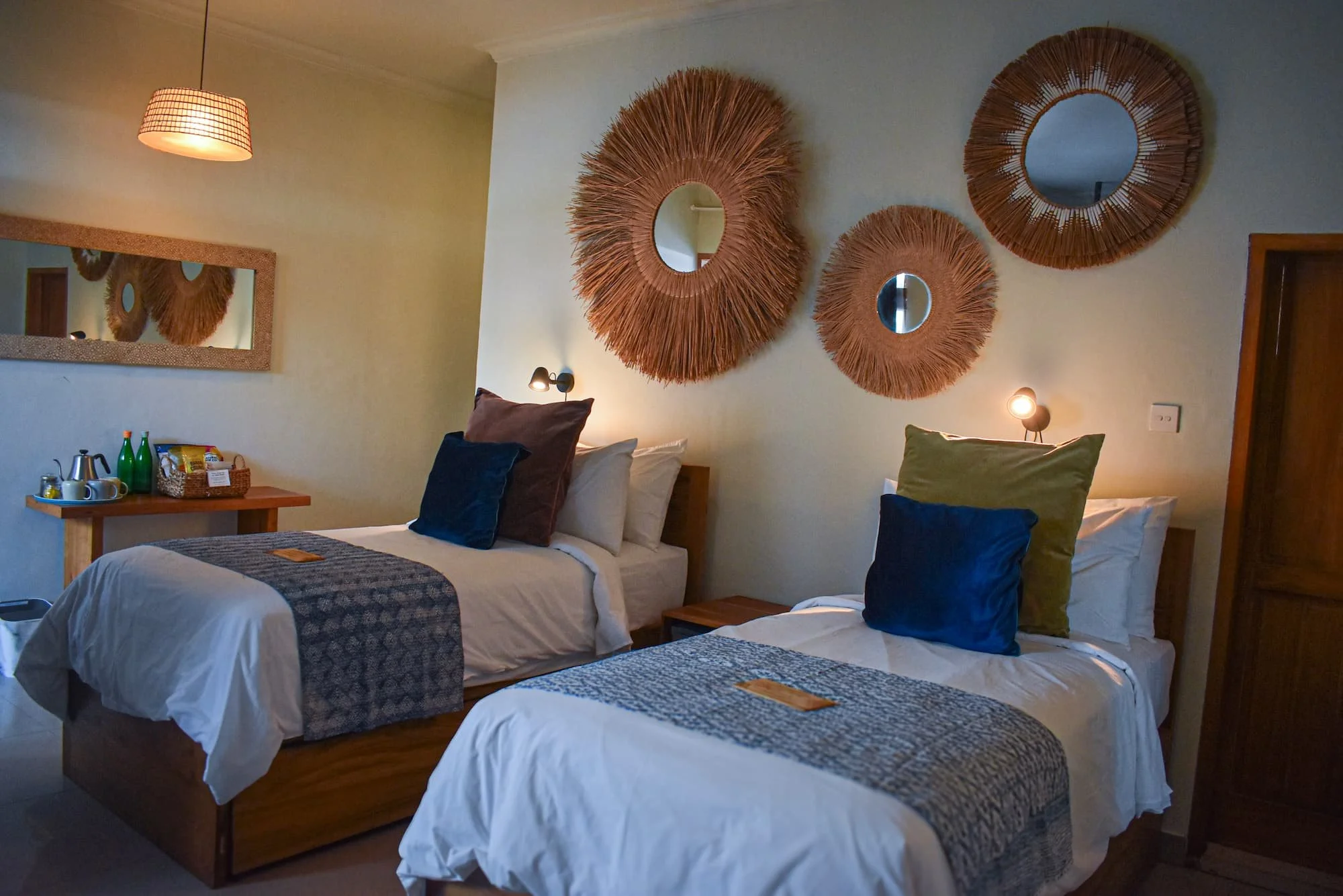 A bedroom with three beds, each decorated with pillows and a blanket, inside a cozy room with wall-mounted lamps. The wall features three round mirrors with woven rattan frames. There is a wooden nightstand with a tray of beverages and snacks. A larg