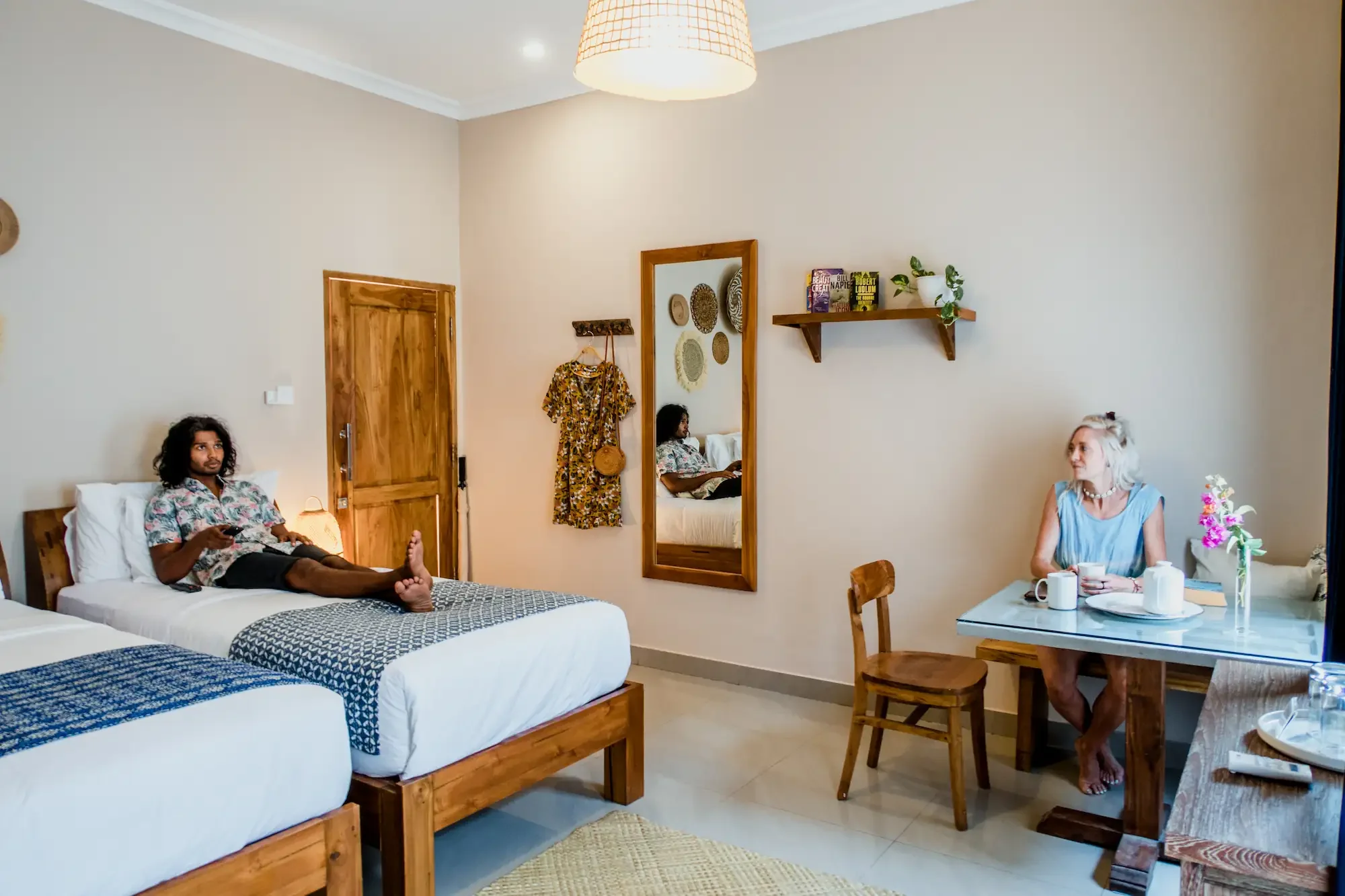 A cozy hotel room with two beds, one occupied by a man using a phone, and the other by a woman sitting at a dining table with coffee cups. The room features wooden furniture, a mirror, wall decorations, and a ceiling light.