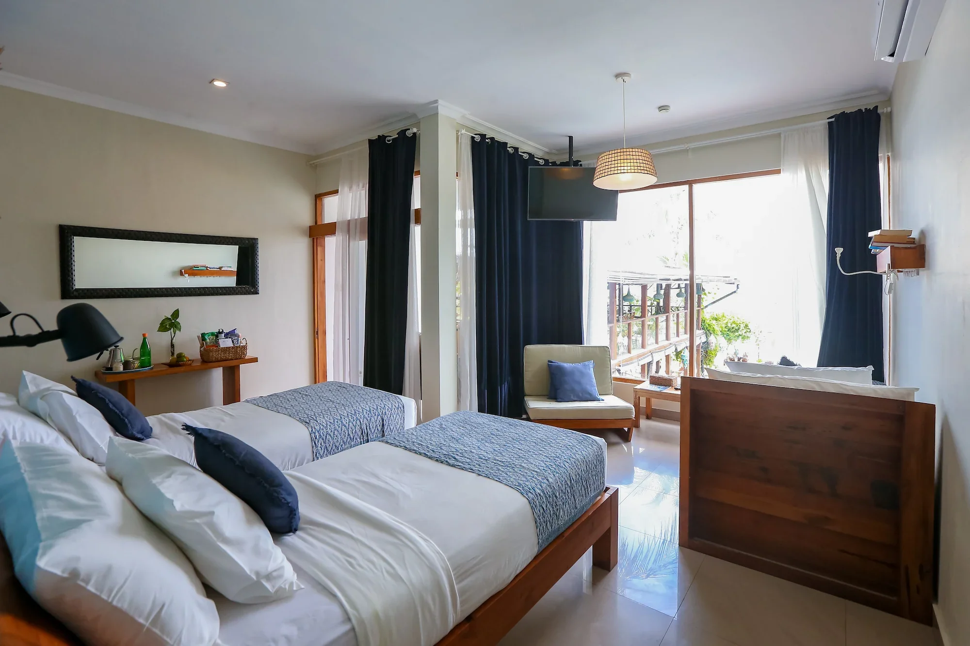 Bright hotel room with two single beds, a small wooden table, a cream armchair, large windows with white and black curtains, a wall-mounted television, and a balcony outside with plants.