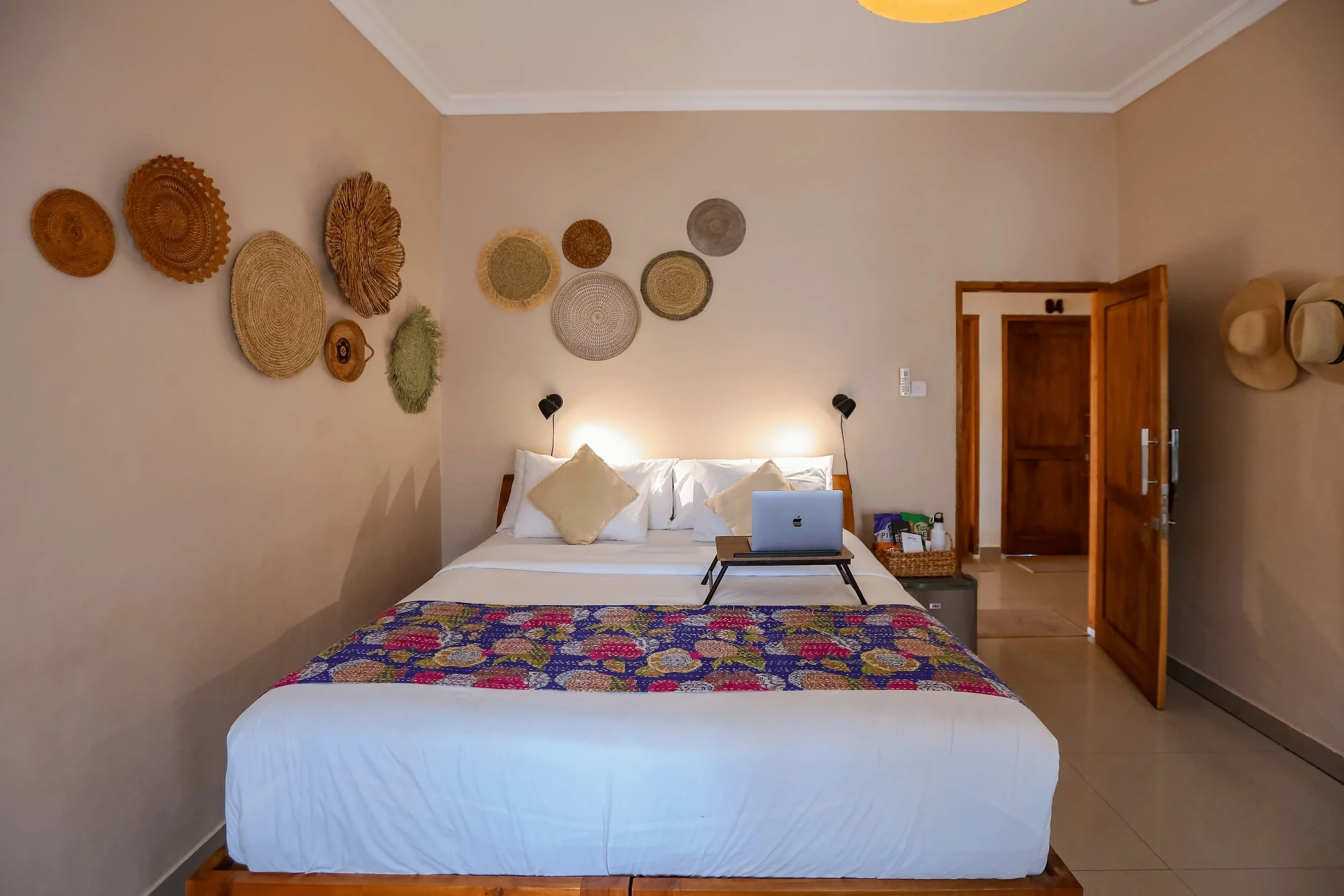 A cozy bedroom with a large bed covered in white linens and colorful quilt, flanked by two black wall lamps. Decorative woven wall hangings are arranged above the bed, and there is a laptop on a small black tray on the bed. The room features beige wa