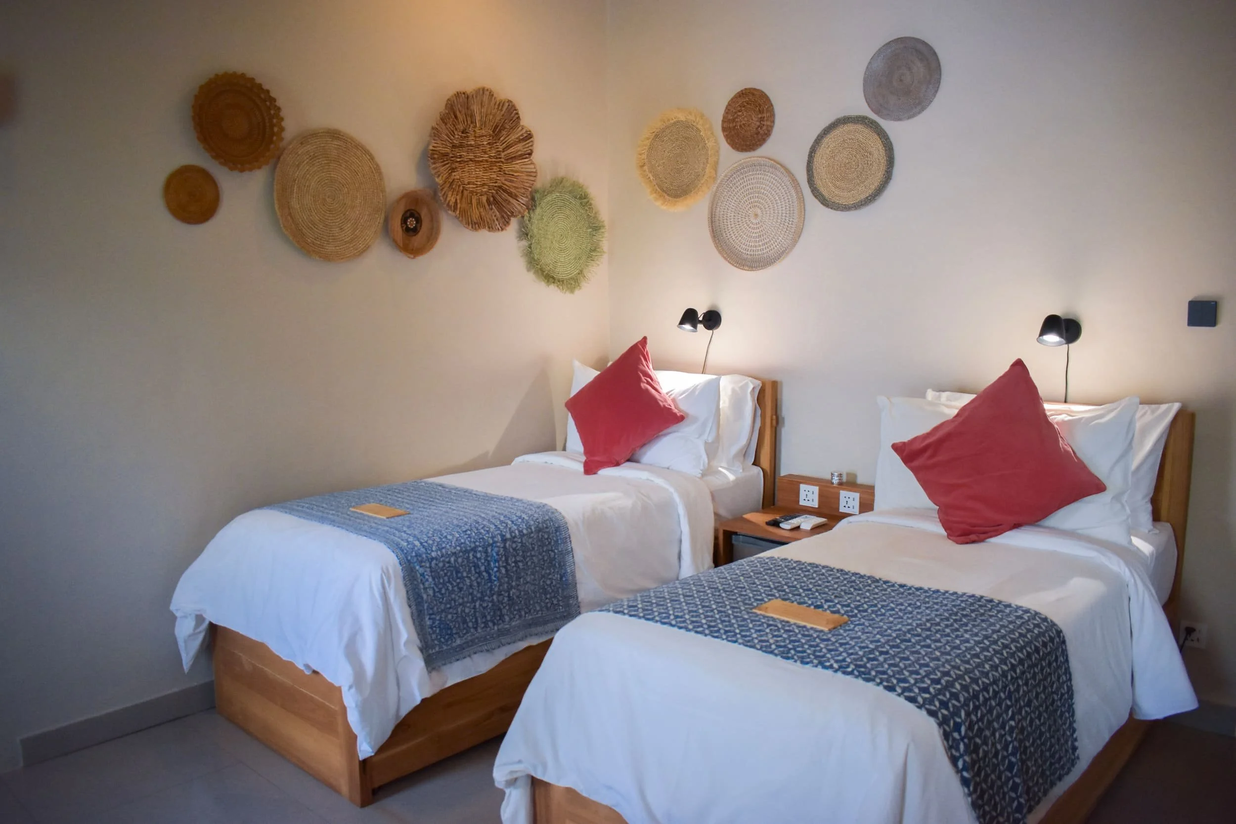 A hotel room with two beds, each with a white bedspread, a red pillow, and a blue patterned bed runner. Above the beds, there is a decorative wall hanging made of woven baskets. Next to each bed, there is a small wooden nightstand with a telephone, a