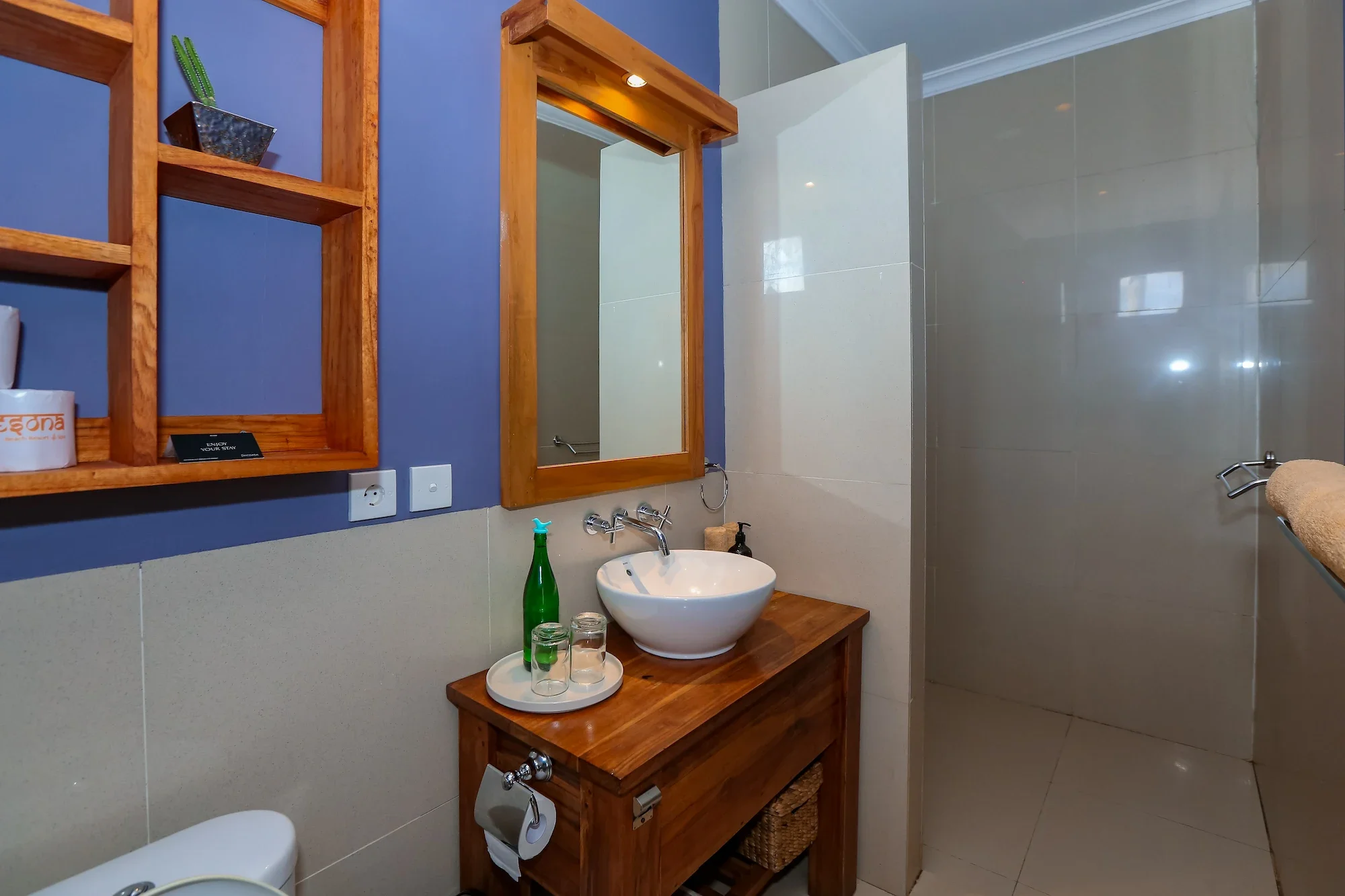 A bathroom with a wooden framed mirror, open wooden shelving on a blue wall, a white vessel sink on a wooden vanity, a green glass bottle, two glasses, a soap dispenser, a toilet, and a shower area with tiled walls.
