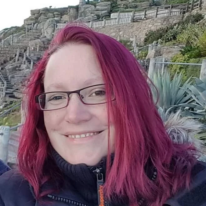 A woman with pink hair and glasses smiling outdoors near rocks and plants.