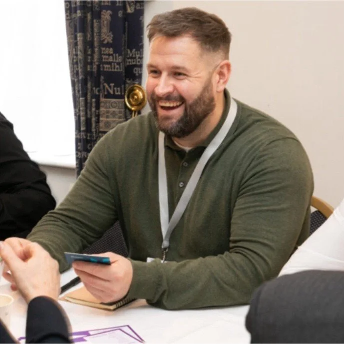 A man with short light brown hair and a beard, wearing a green long-sleeve shirt and a lanyard, is sitting at a table, smiling and holding a smartphone in his right hand. He is in a room with patterned curtains.