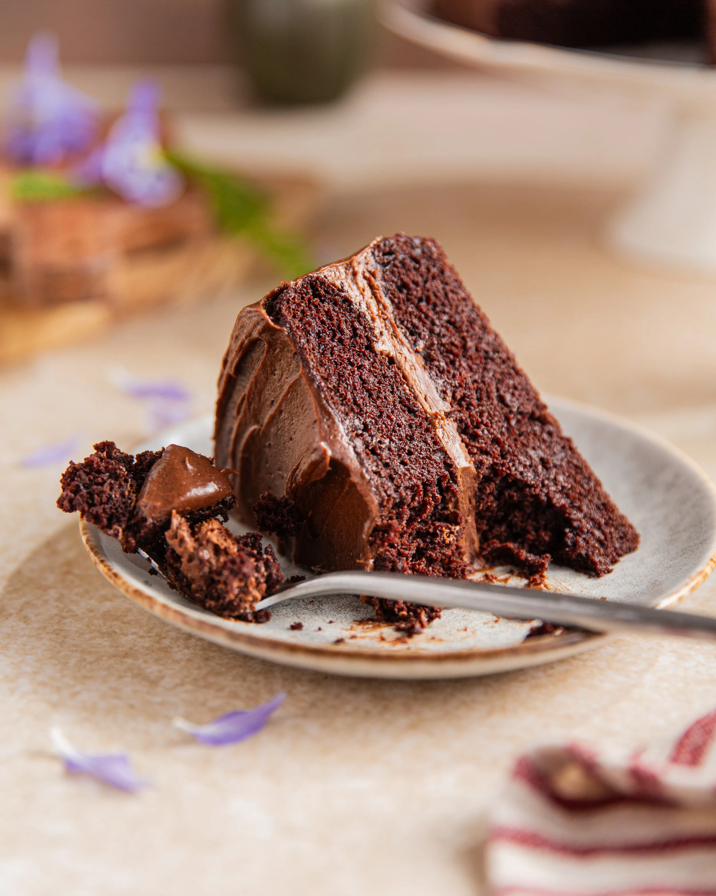 A slice of chocolate cake on a plate with a fork, with purple and red flower petals scattered on a beige surface in the background.