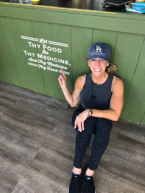 Anne-Marie Berte with a baseball cap, sitting on the floor, smiling, pointing to a green wall with a white quote that says 'Let thy food be thy medicine, and thy medicine thy food.'