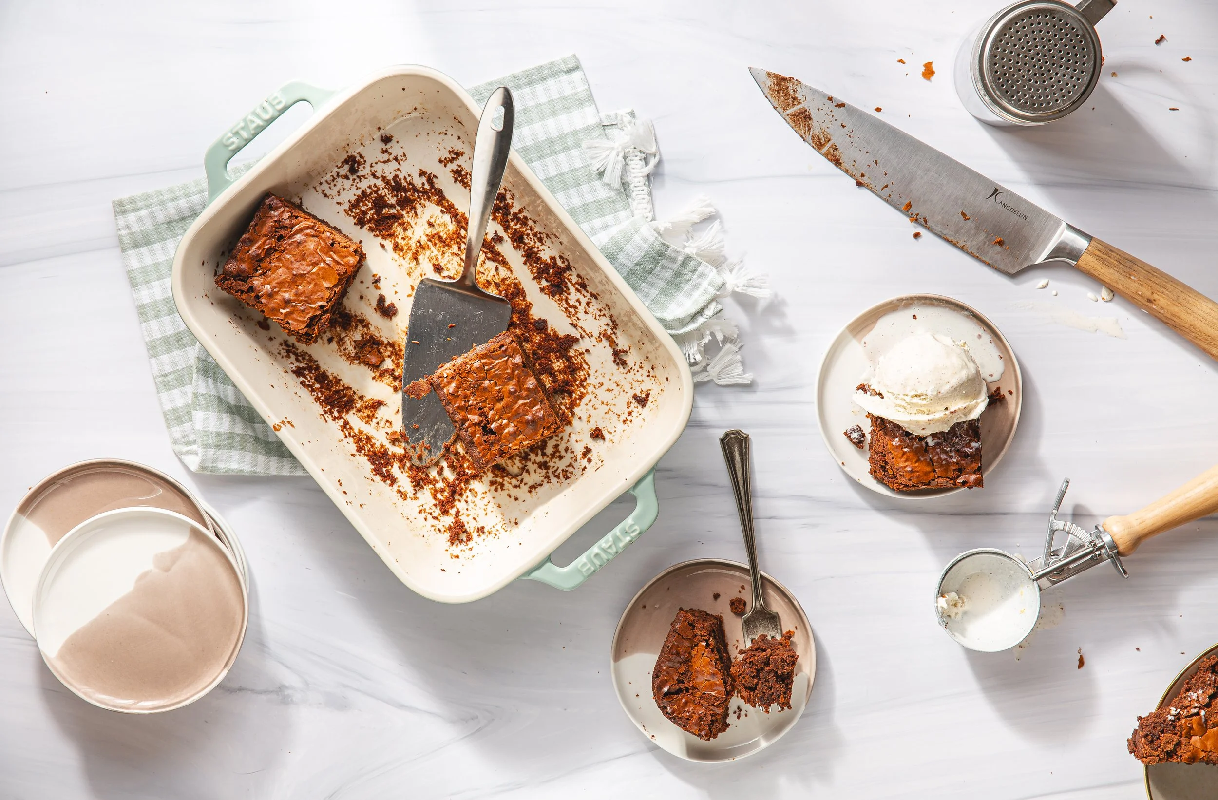 Empty baking dish with two pieces of chocolate cake, surrounded by cream, chocolate, and baking tools on a white surface.