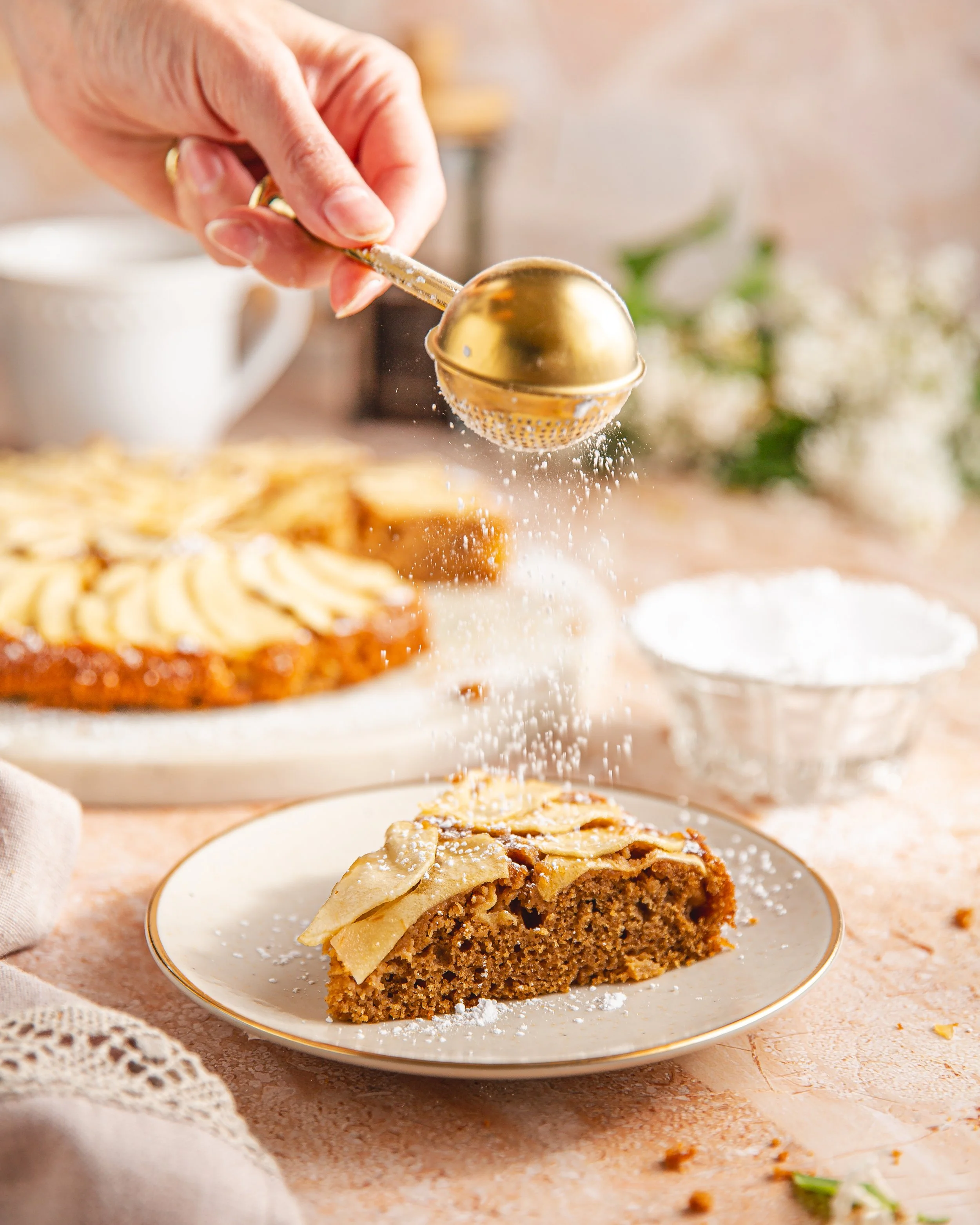 Hand sprinkling powdered sugar on a slice of homemade apple pie on a plate, with additional slices of pie in the background and a cup on a table.