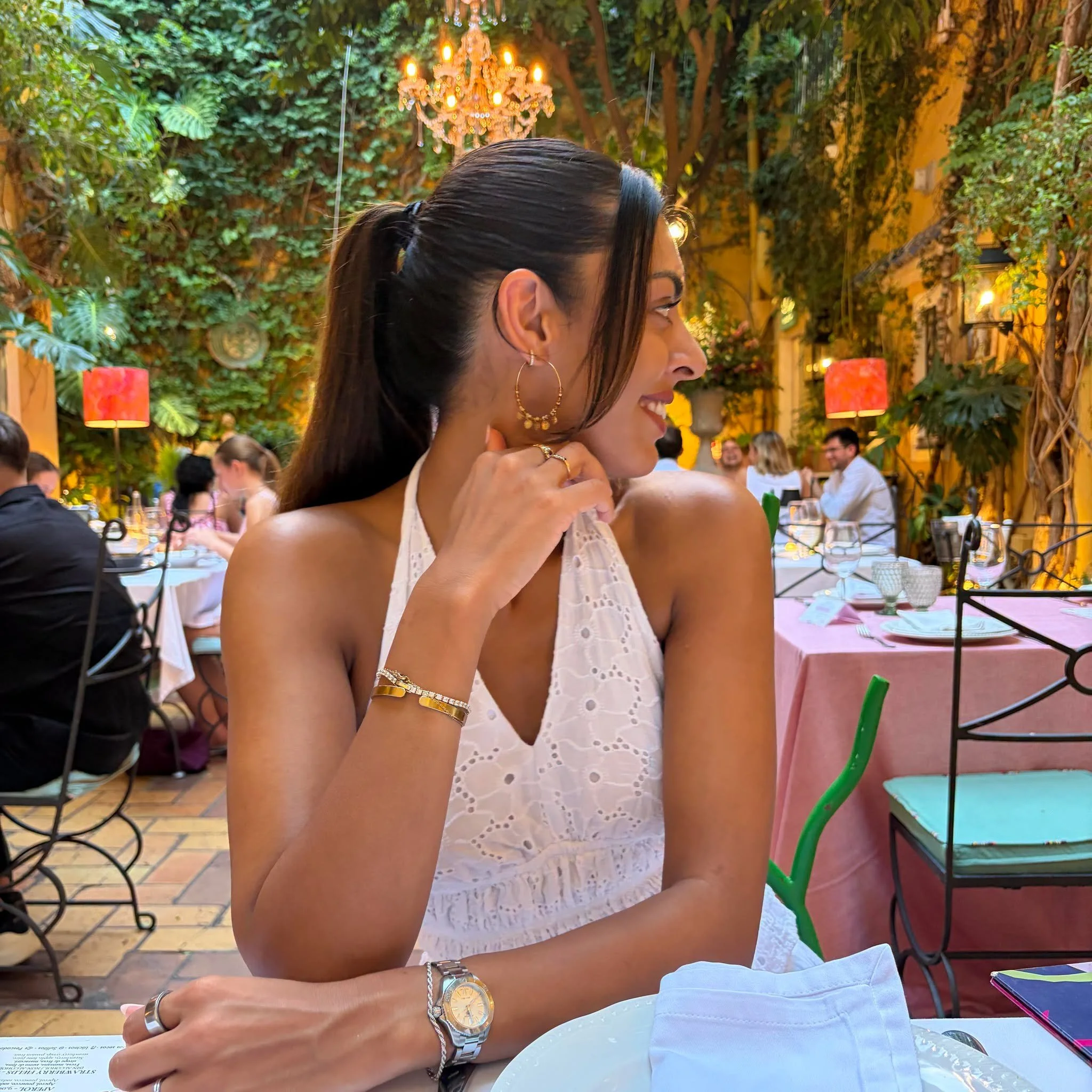 A woman with long dark hair tied in a ponytail, wearing earrings, a watch, and jewelry, smiling at a restaurant table with pink tablecloths, set with wine glasses, plates, and cutlery. The restaurant has a lush green interior with a chandelier hanging from the ceiling, and other diners can be seen in the background.
