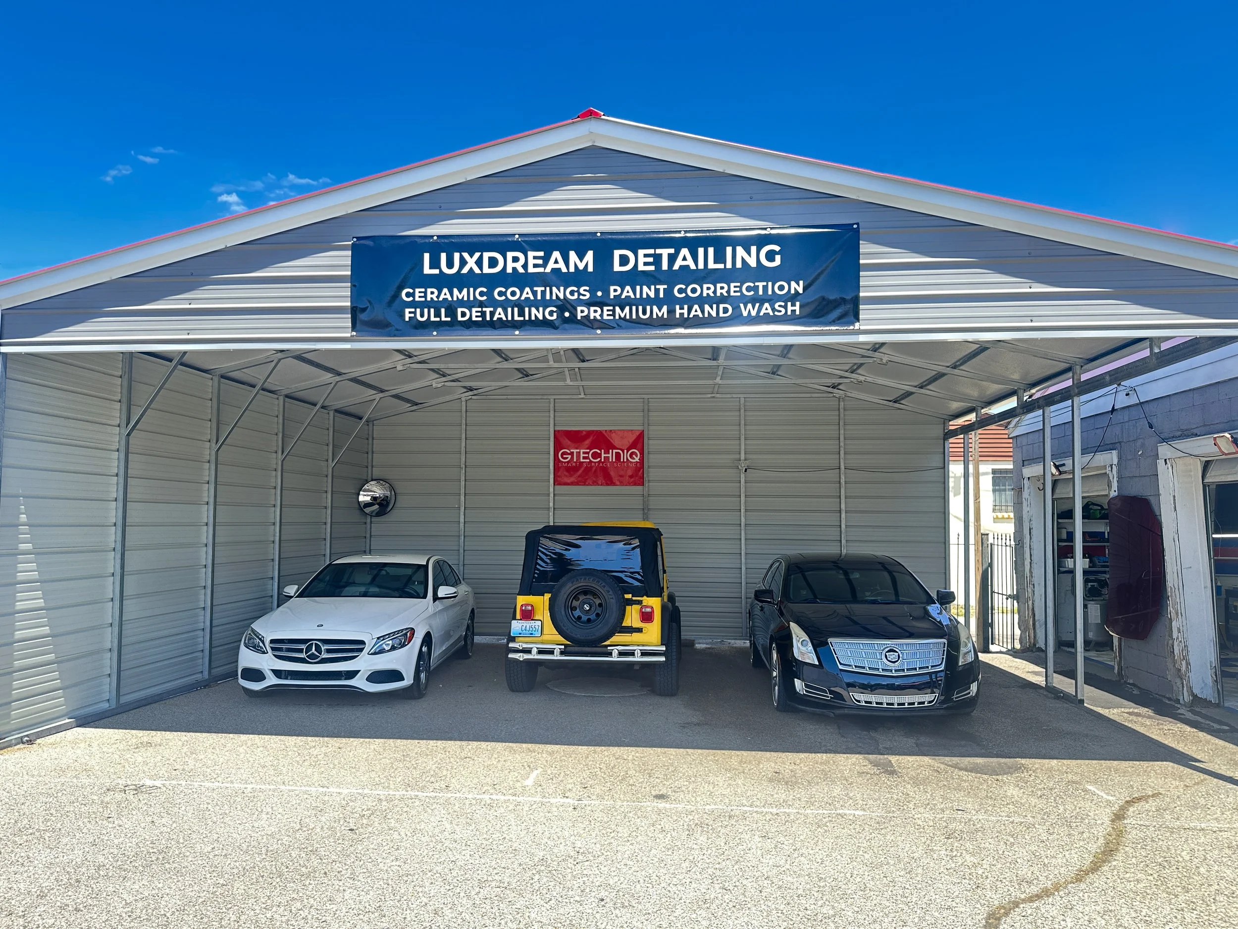 A car detailing garage with a blue sign reading 'Luxdream Detailing' and services like ceramic coatings, paint correction, full detailing, and premium hand wash. Inside, there are three parked cars, a white Mercedes, a yellow Jeep with a spare tire, and a black Cadillac, under a metal roof structure.