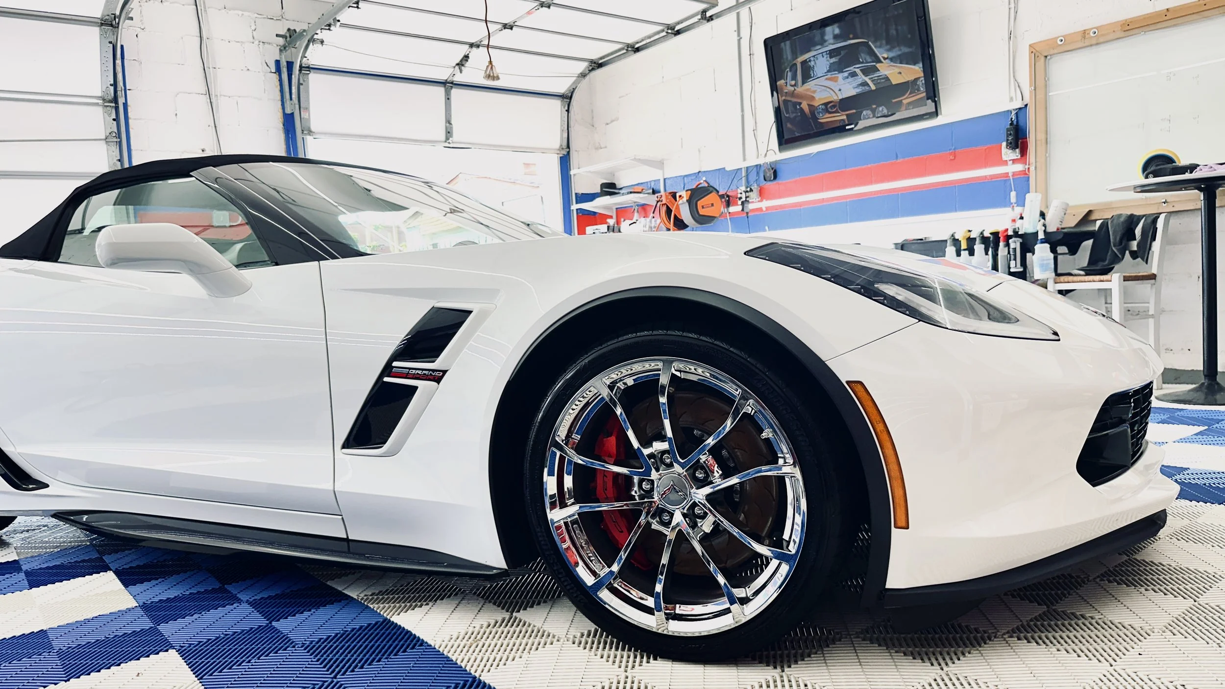 White Chevrolet Corvette with black convertible top inside a garage or detailing shop, with tools and a TV showing a similar car in the background.