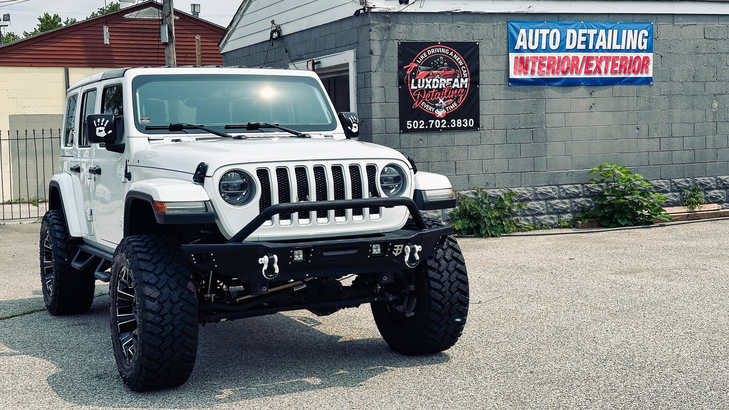 A white Jeep with large off-road tires and a black front bumper parked in front of a gray building with a sign that reads 'Auto Detailing Interior/Exterior' and another sign for Luxdream Detailing, with a phone number. There are green plants growing 