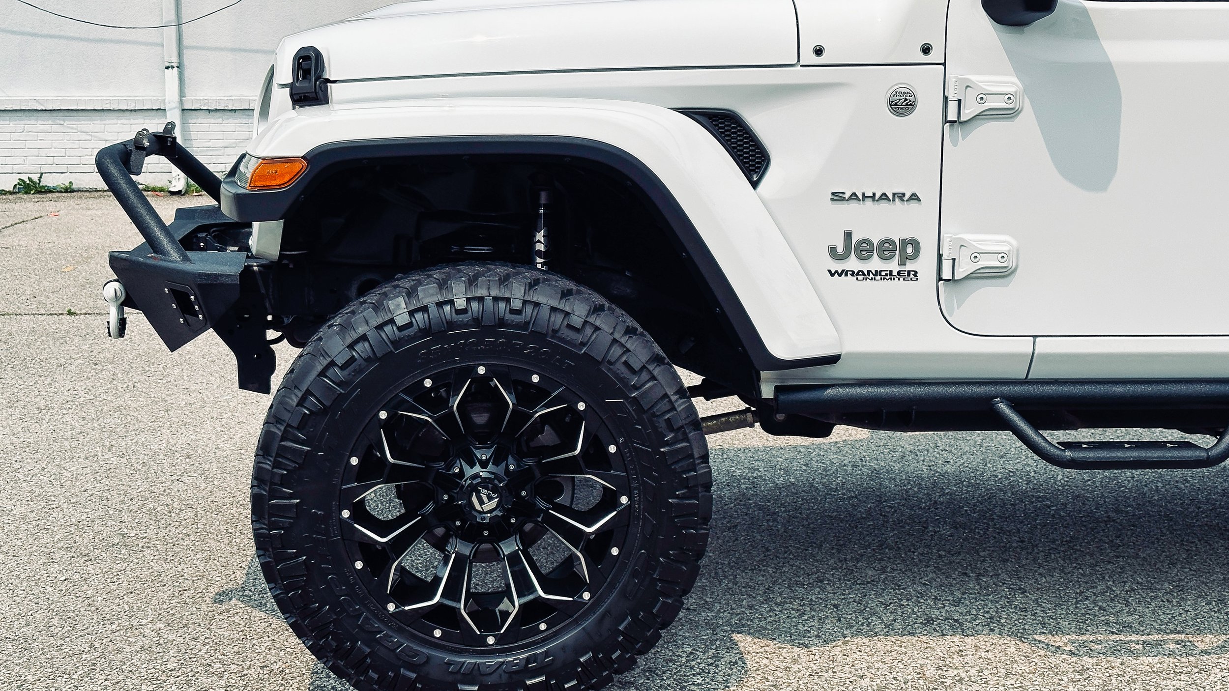 Close-up of the front part of a white Jeep Sahara Unlimited vehicle, showing the large tire, fender, and front bumper with off-road features.
