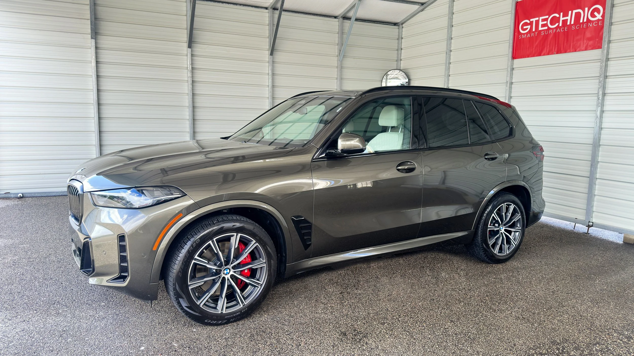 A metallic gray BMW SUV parked inside a covered car storage area with gray walls and a red sign that reads 'GTECHNIQ SMART SURFACE SCIENCE'.
