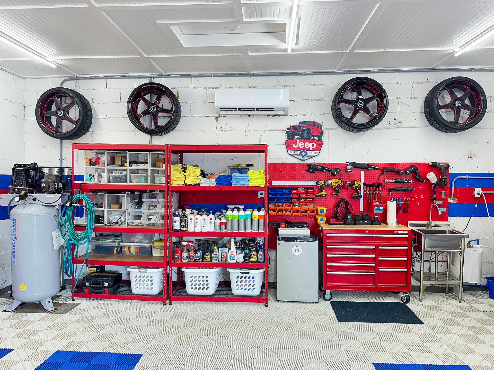 Workshop with wall-mounted wheels, red tool chest, workbench, pegboard with tools, shelves with bottles and supplies, air compressor, and cleaning supplies in front of a white brick wall.