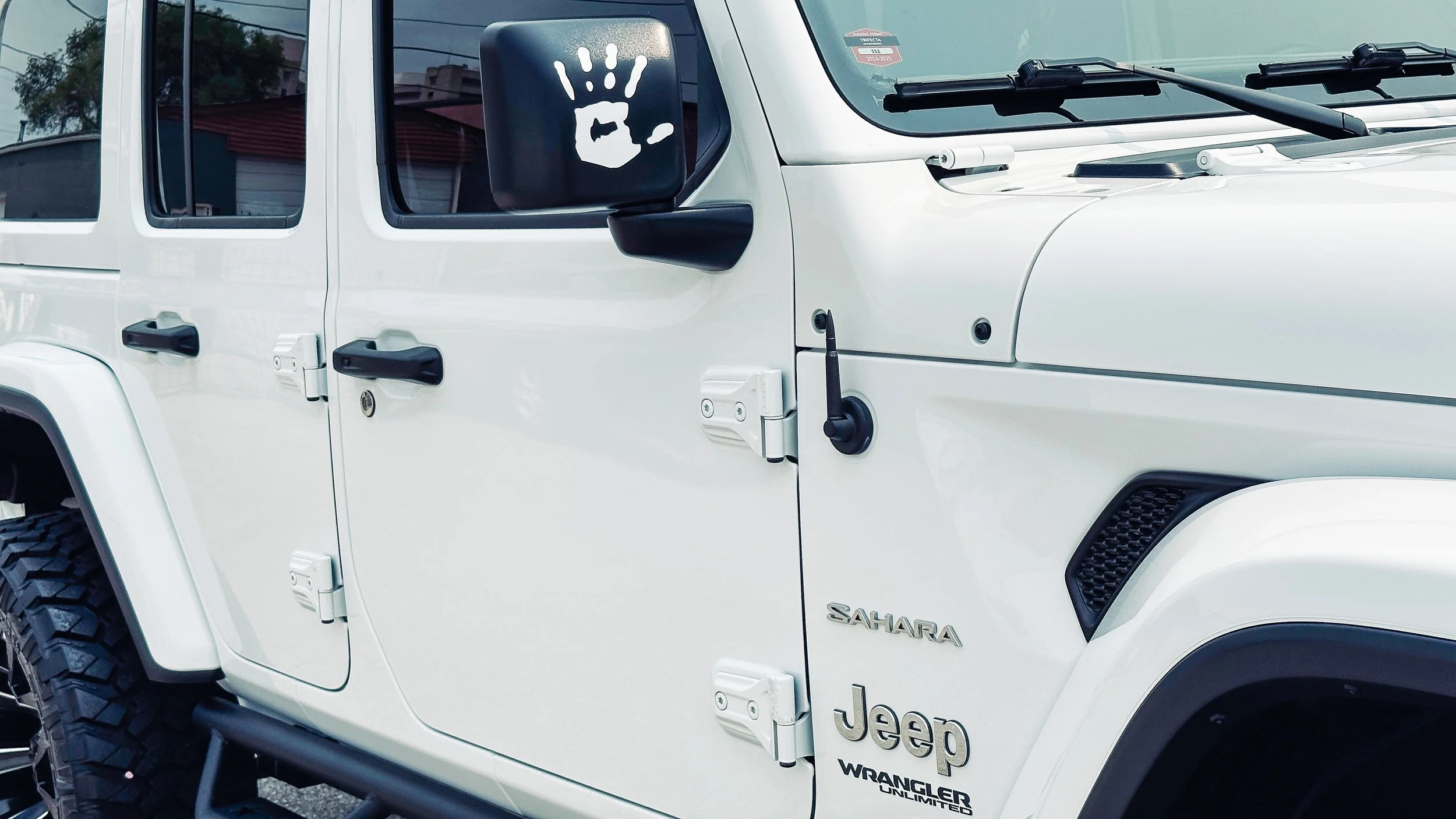 A white Jeep Sahara vehicle with an added decal of a black box featuring a white hand giving an okay gesture. The vehicle has black side mirrors and door handles, and various model badges including 'Sahara' and 'Wrangler Unlimited' on the front fende