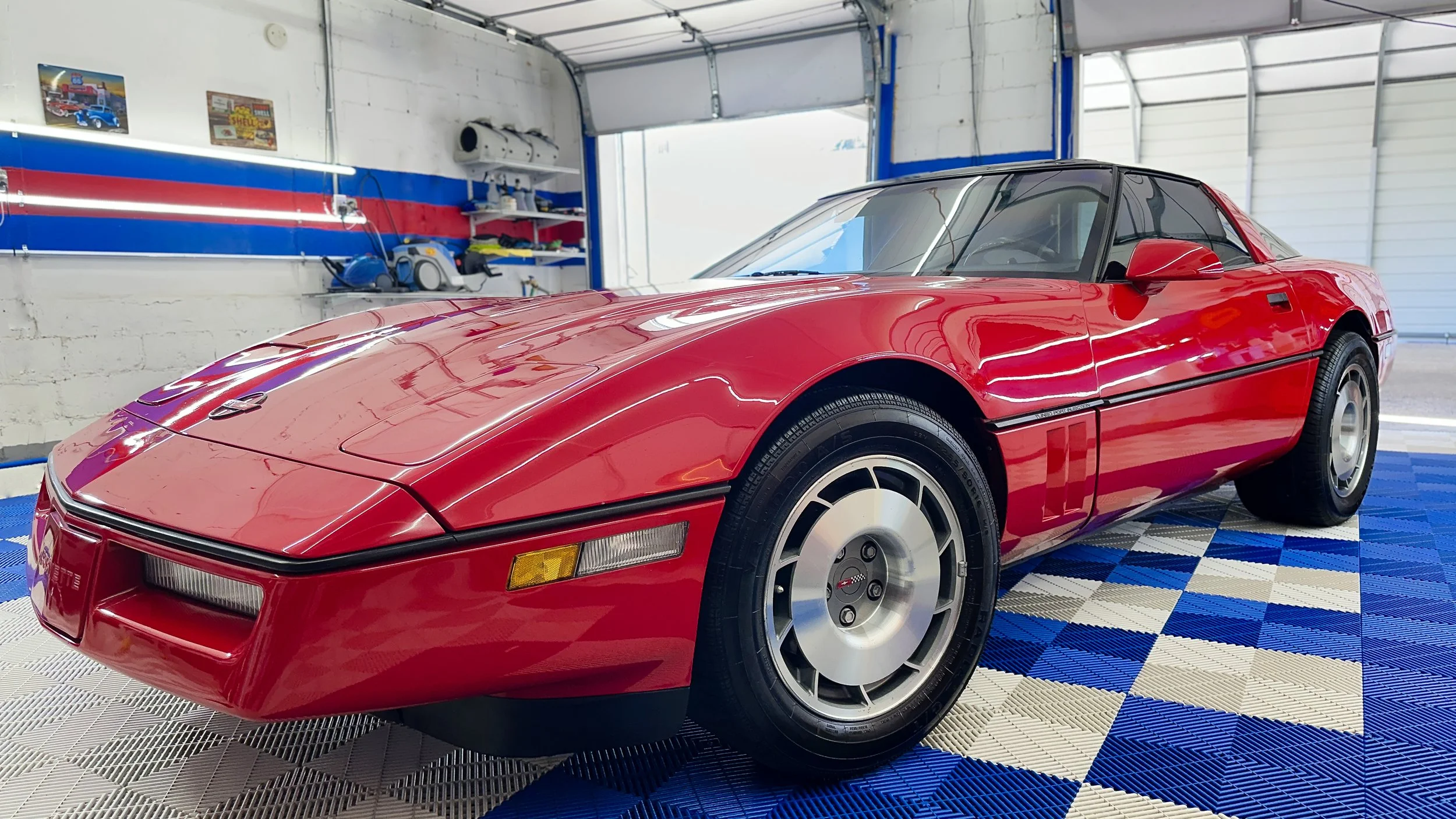 A red sports car is parked inside a garage with white and blue checkered flooring and white brick walls. The garage has some tools and posters on the wall.