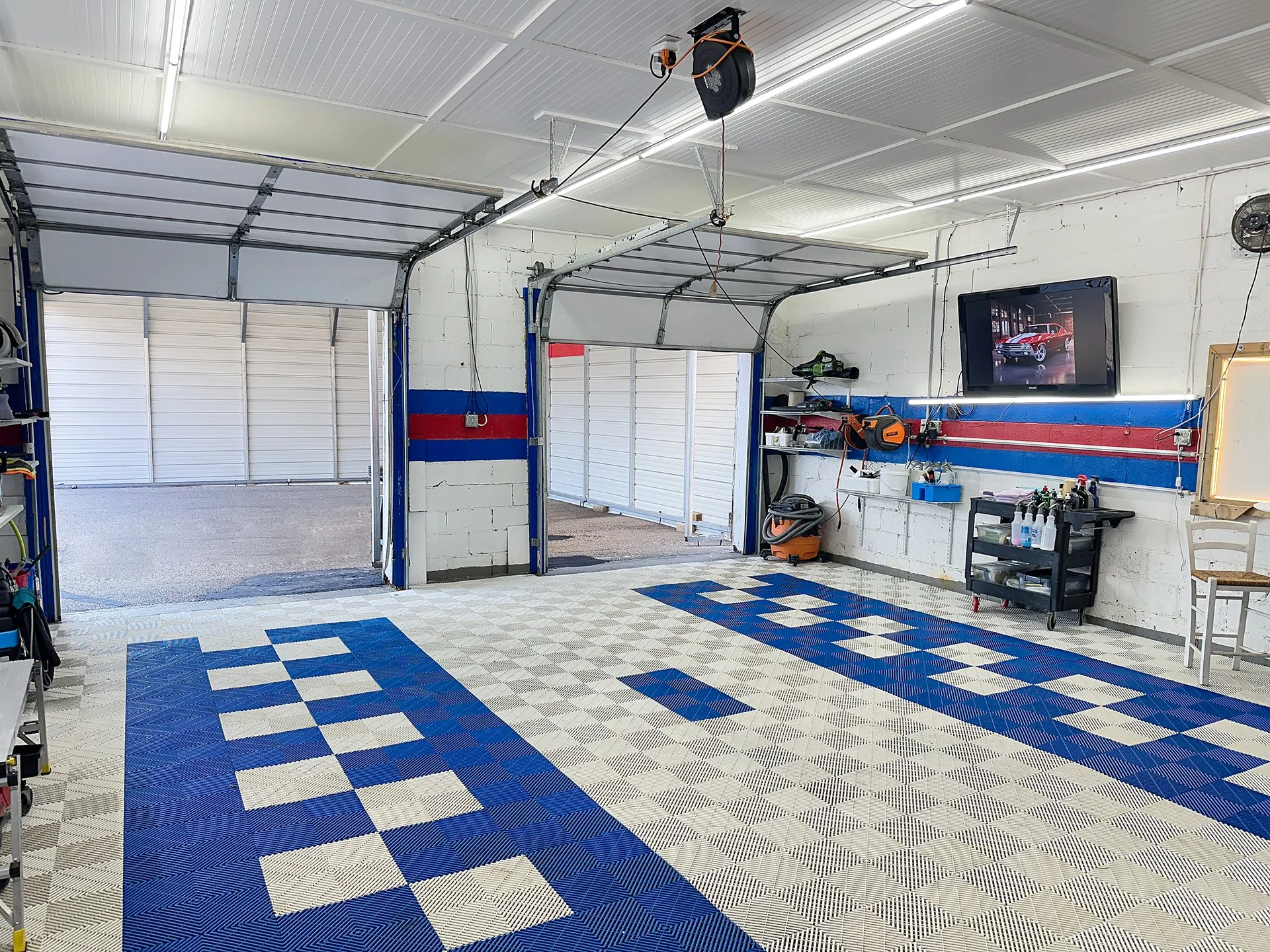 Clean garage with blue, white checkered flooring, white brick walls, two open garage doors, and a wall-mounted TV showing a classic red car. Tools and cleaning supplies are organized on shelves and carts.