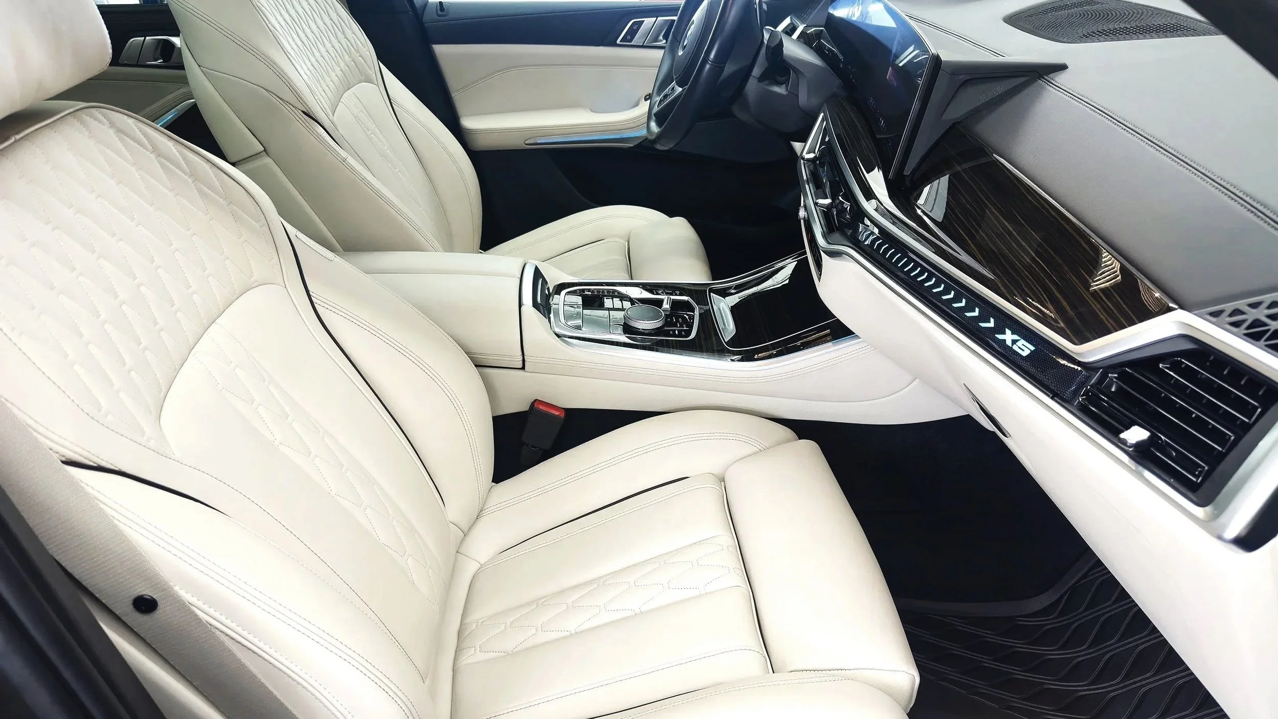The interior of a luxury car with cream-colored quilted leather seats, a sleek black center console with control knob and buttons, a digital dashboard displaying 'X5', and a dashboard with wood and metallic accents.
