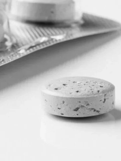 A close-up of a round, speckled pill tablet on a white surface with a partially visible blister pack and a container in the background.