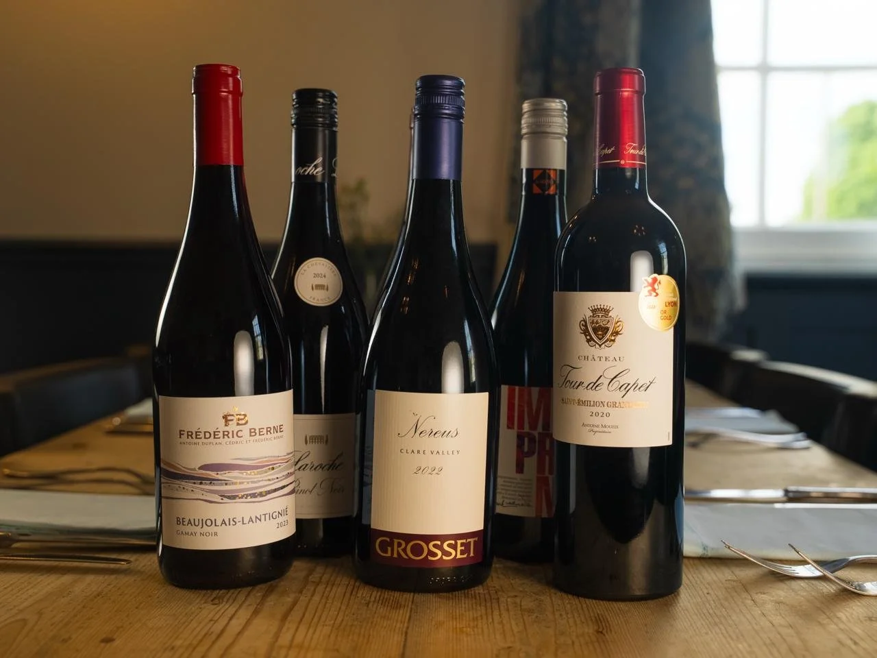 Five wine bottles arranged on a dining table with table settings, in a room with a window and curtains in the background.