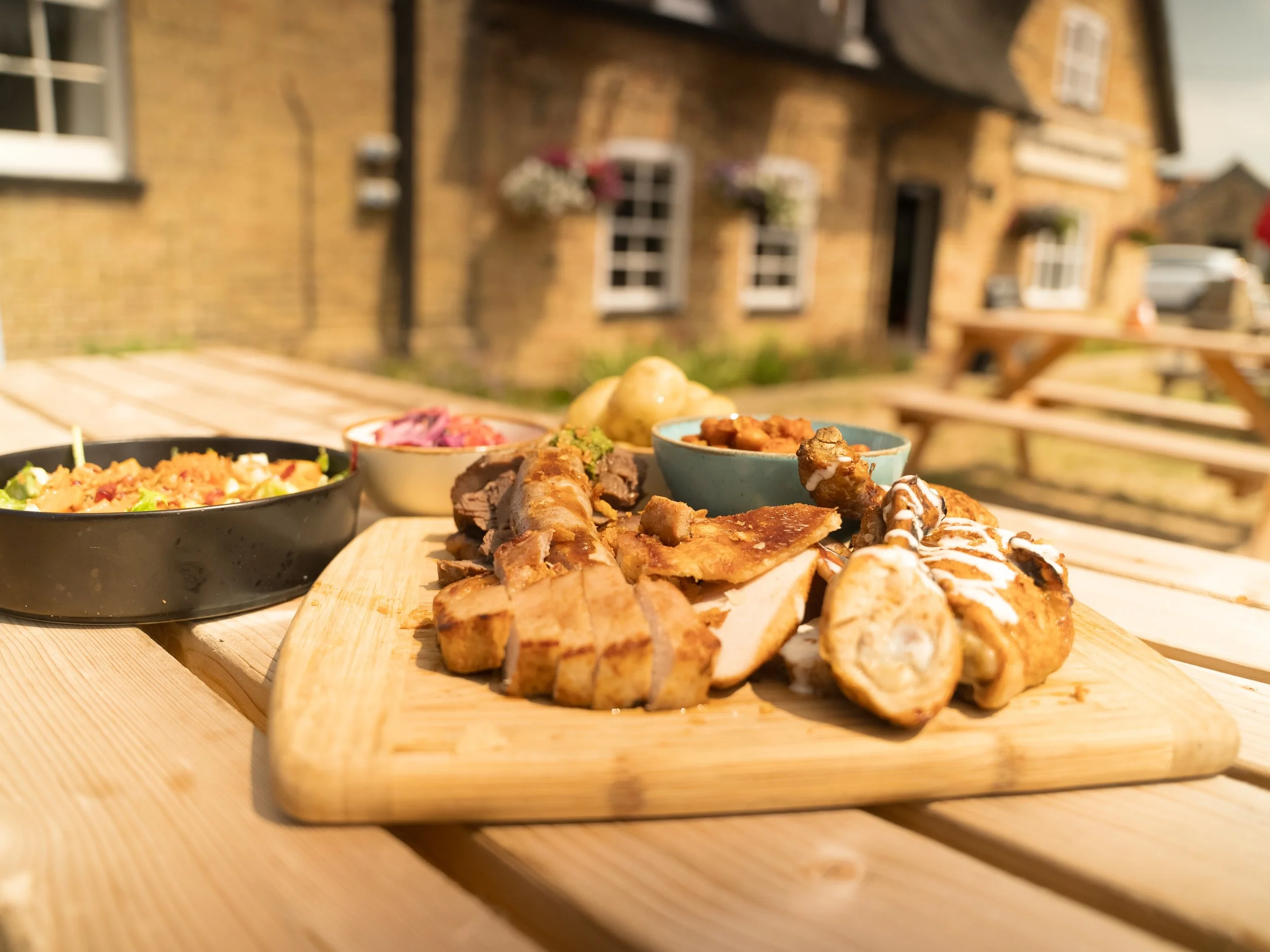 A variety of dishes including grilled meats, salads, and side dishes served on a wooden tray outdoors with a brick building and a wooden picnic table in the background.